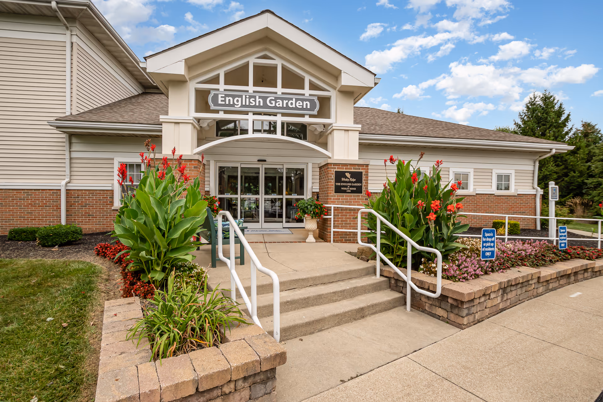Entrance to a building labeled English Garden at Wesley Ridge, featuring a ramp and stairs with white railings, surrounded by landscaped flower beds with red flowers and greenery under a partly cloudy sky.