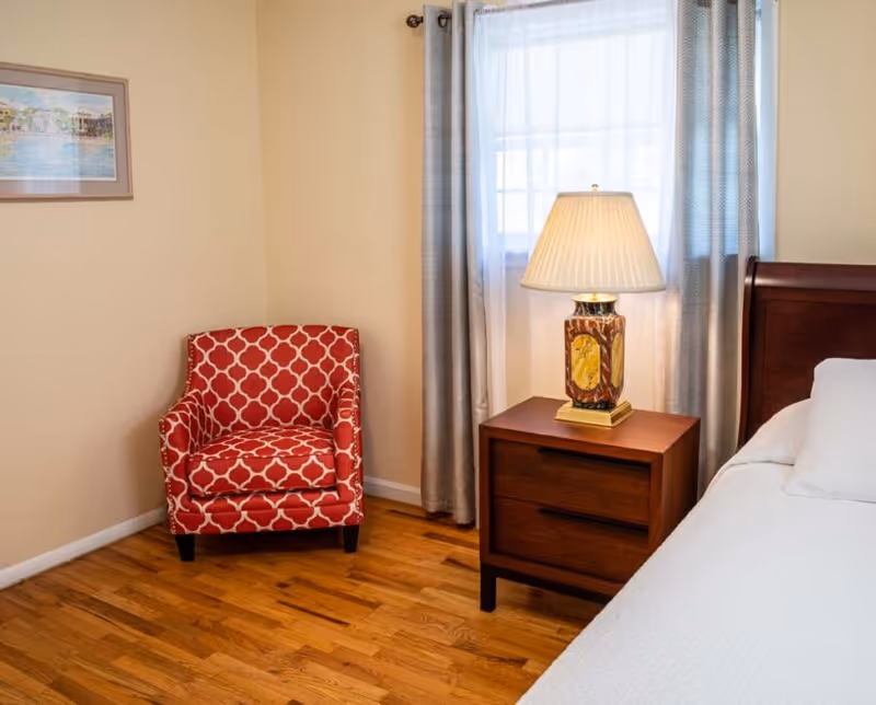 A cozy bedroom corner with a red patterned chair, nightstand, lamp, and window with curtains.