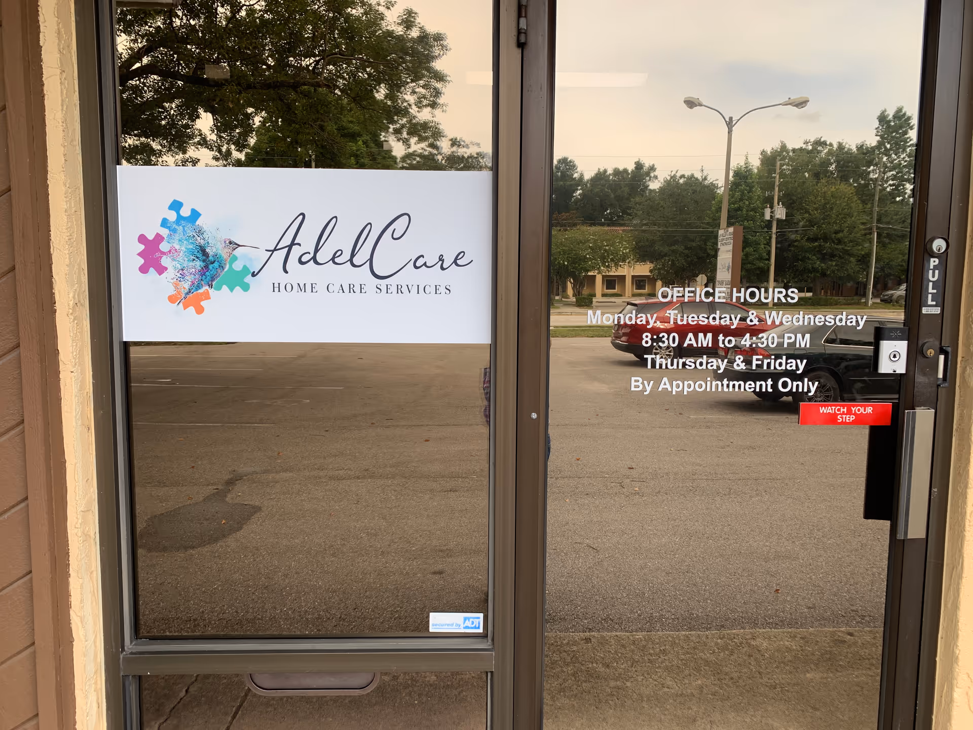Glass entrance door displays AdelCare Home Care Services sign; exterior view with parking lot outside.