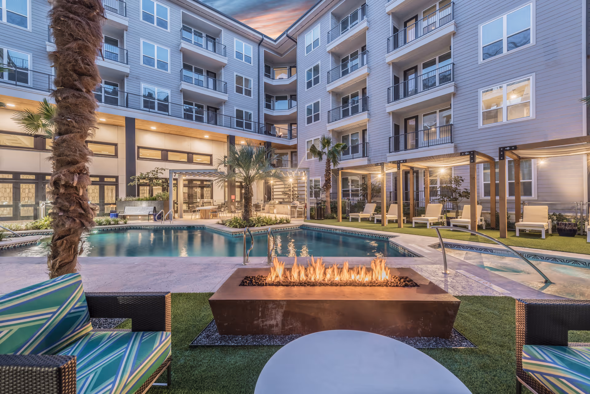 Outdoor courtyard area of a senior living facility featuring a swimming pool, a modern rectangular fire pit with flames, palm trees, lounge chairs under wooden pergolas, and a multi-story building with balconies in the background during dusk.