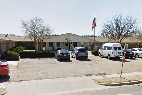 Front exterior view of Lakeridge Nursing & Rehabilitation facility with a parking lot in front, several parked cars, an American flag on a flagpole, and leafless trees on either side of the building entrance.