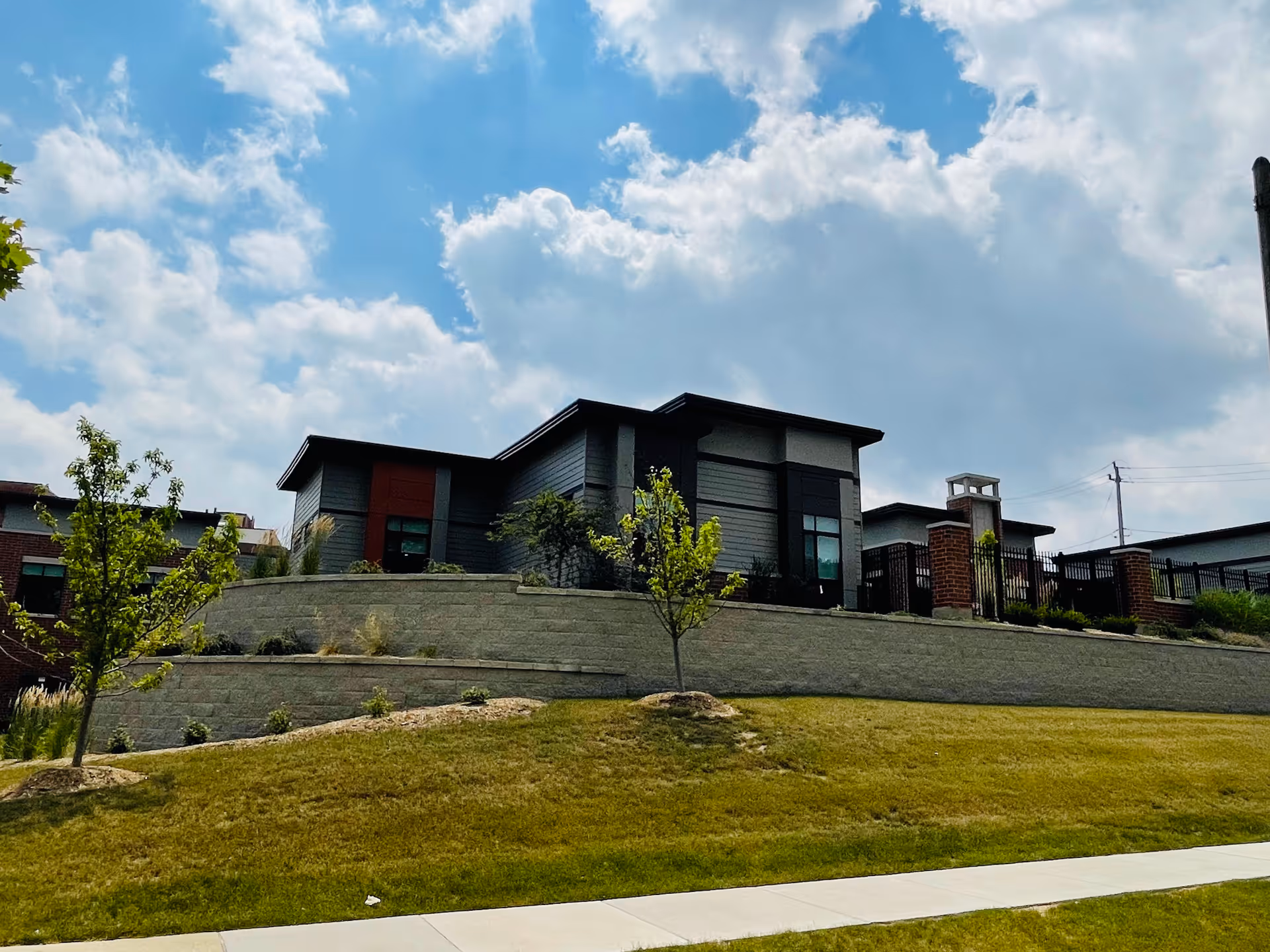 Modern single-story senior living building atop a terraced retaining wall with a grassy slope, small trees, and a cloudy blue sky.