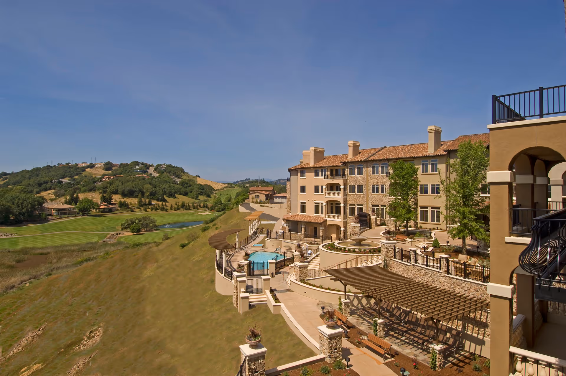 Multi-story Mediterranean-style senior living building with terraces, a pool, pergolas and landscaped courtyard overlooking a golf course and hillside.