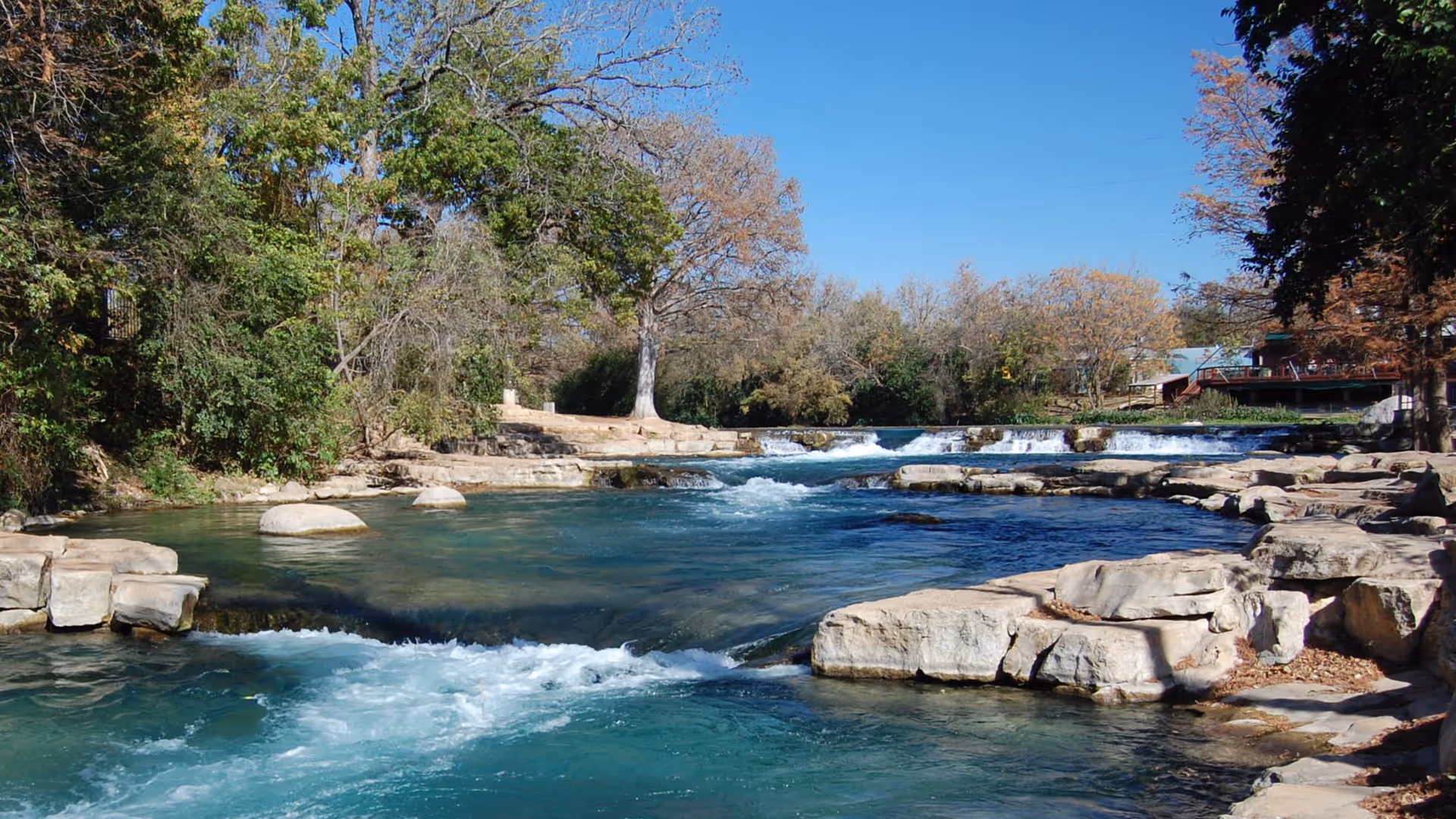 Clear river flows over rocks surrounded by trees under a blue sky.