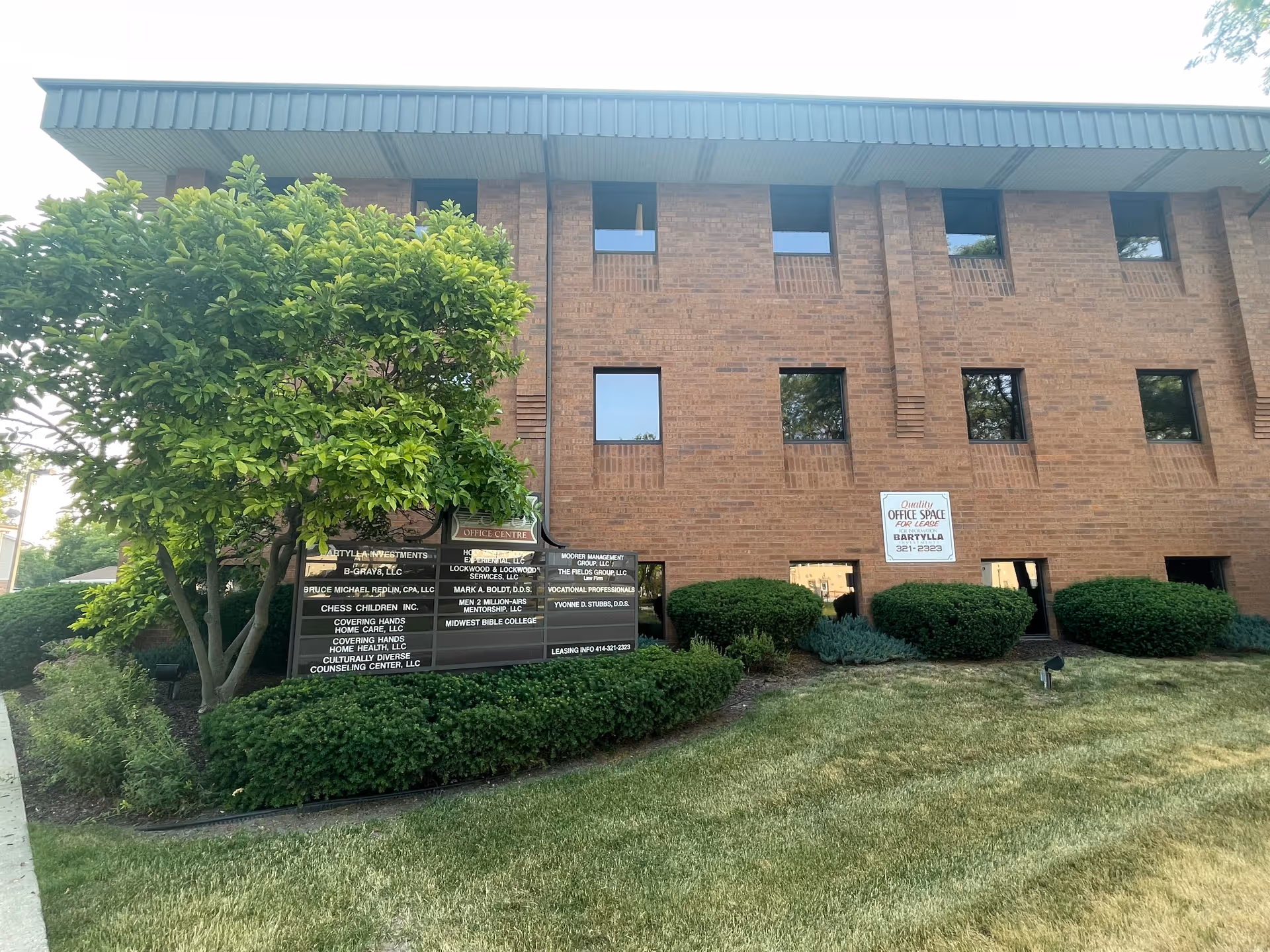 Brick office building with signs, trimmed bushes, and a green lawn in front.