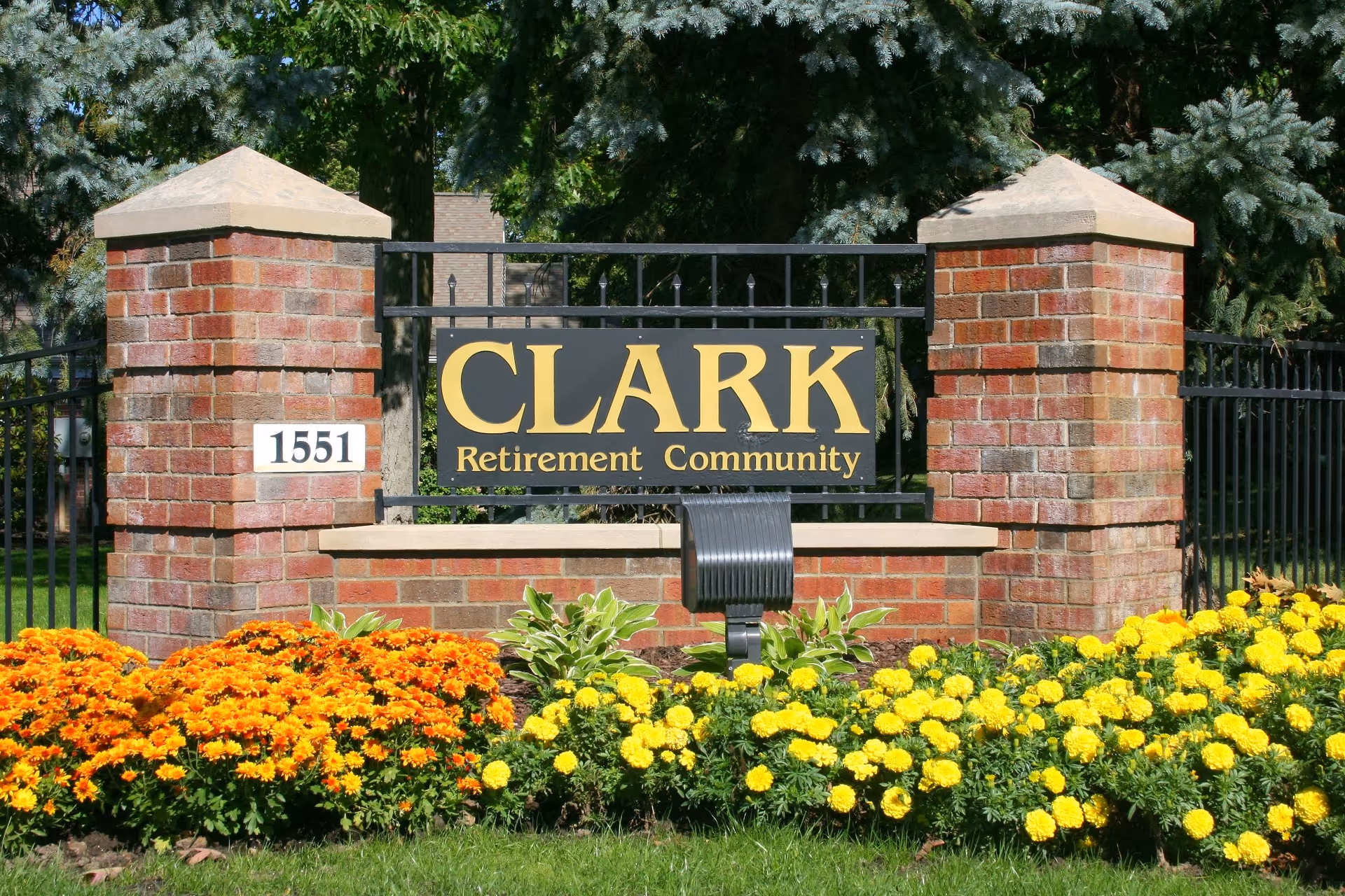 Brick entrance sign reading "CLARK Retirement Community" with the address 1551 and colorful flowerbeds in front.