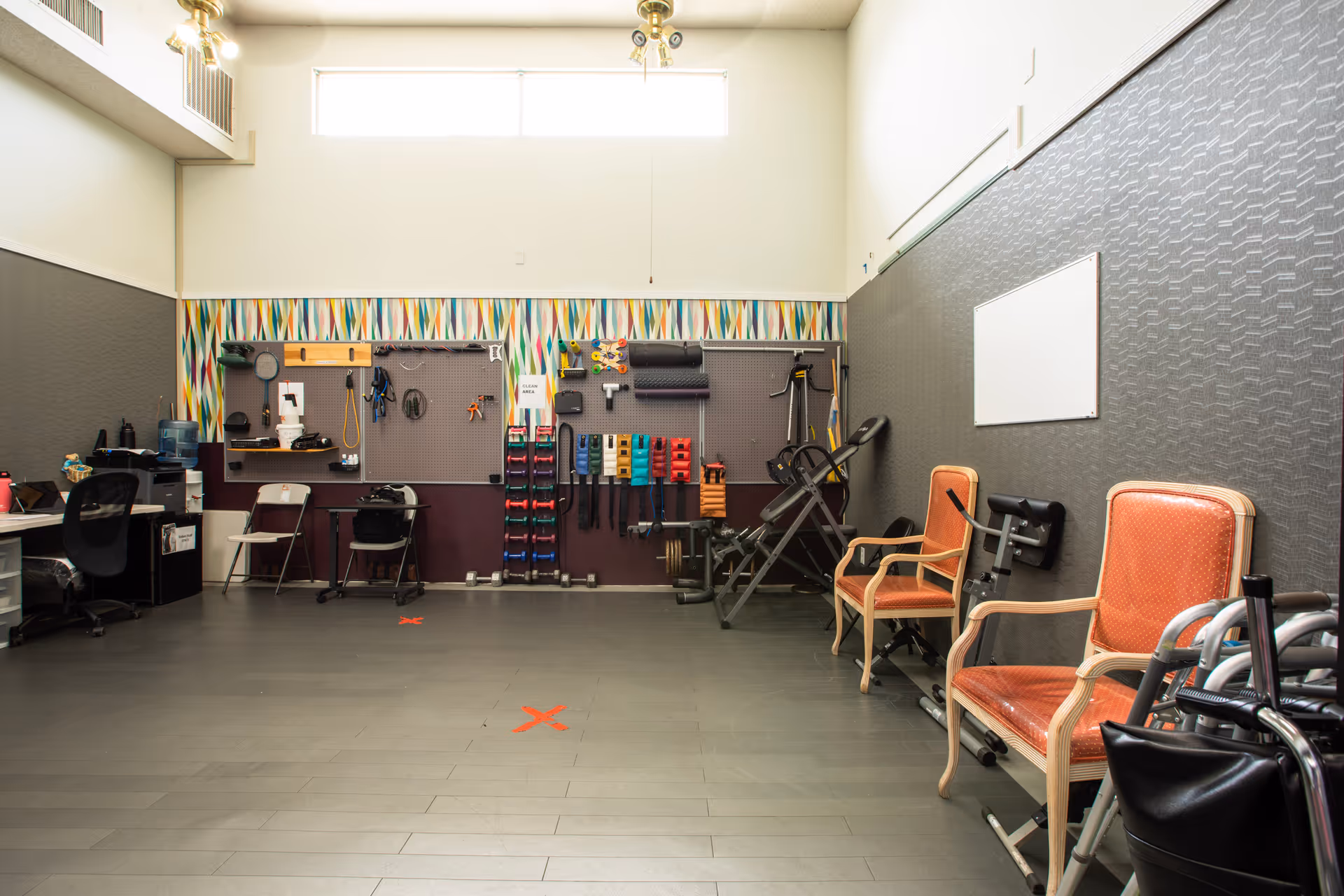 A spacious room with exercise equipment and therapy tools organized on the wall, two orange cushioned chairs, a walker, and a desk with office supplies. The room has high windows letting in natural light and a patterned wallpaper border near the ceiling.