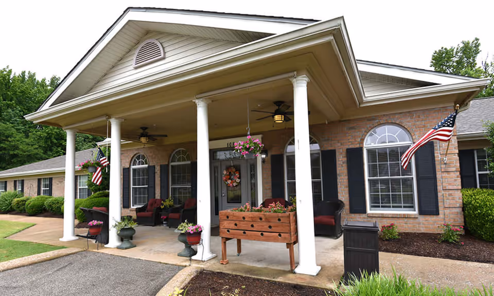 Front exterior view of a single-story brick building with a covered porch supported by white columns. The porch has seating with red cushions, hanging flower baskets, and American flags on either side. The building has arched windows with black shutters and is surrounded by greenery.