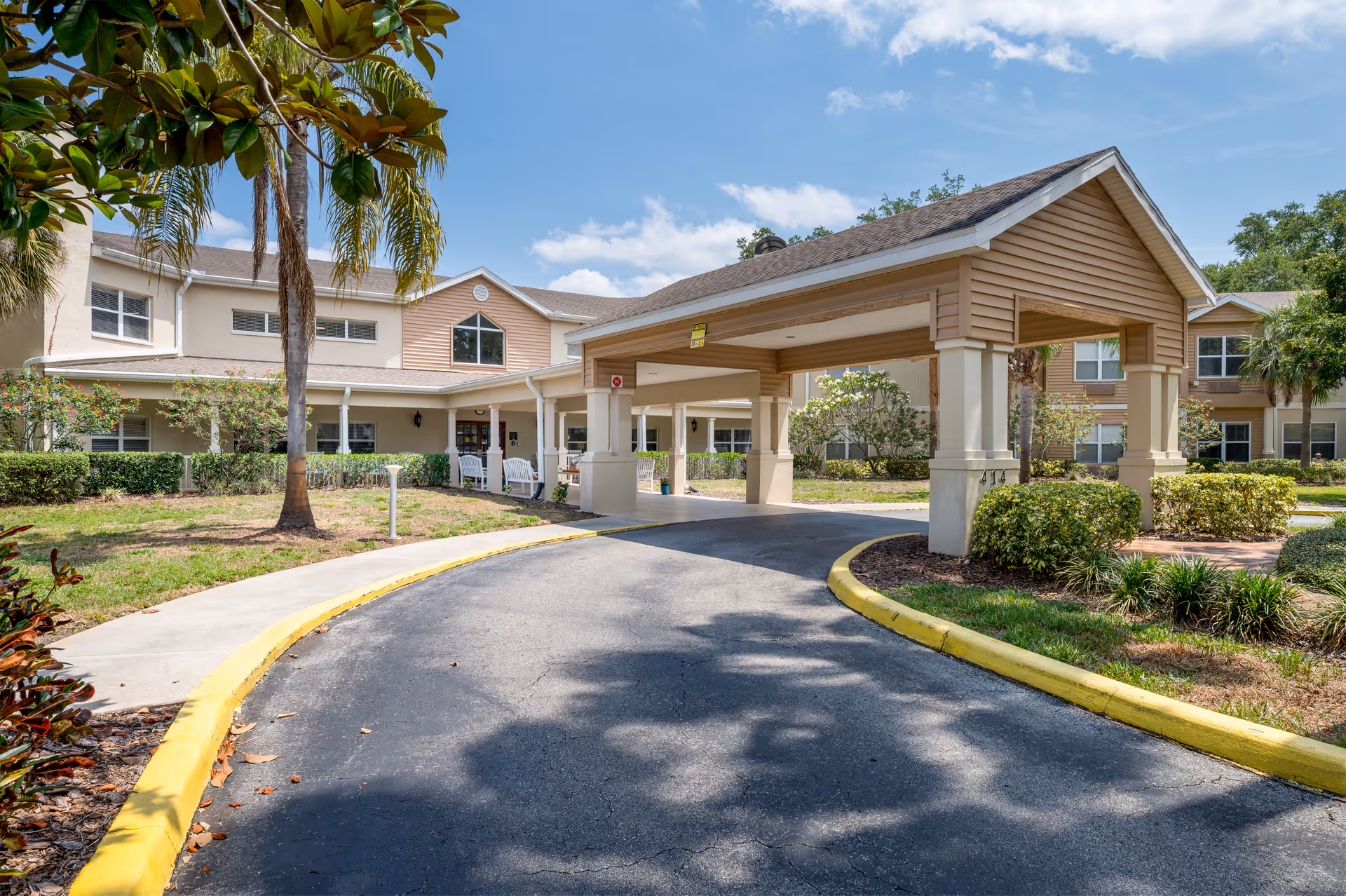 Exterior view of Chapman Manor at Lutz, showing a covered driveway entrance with a peaked roof, surrounded by landscaped greenery and palm trees under a partly cloudy sky.