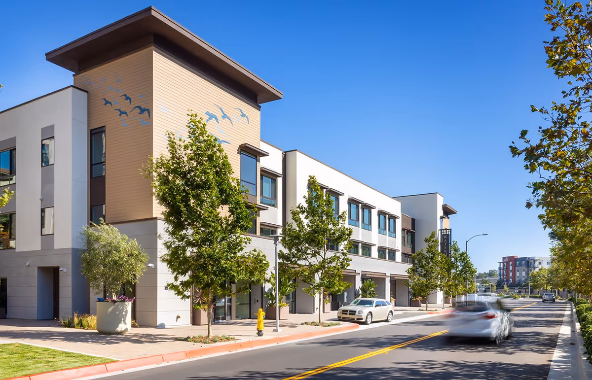 Modern three-story senior living building front on a sunny street with trees and passing cars.