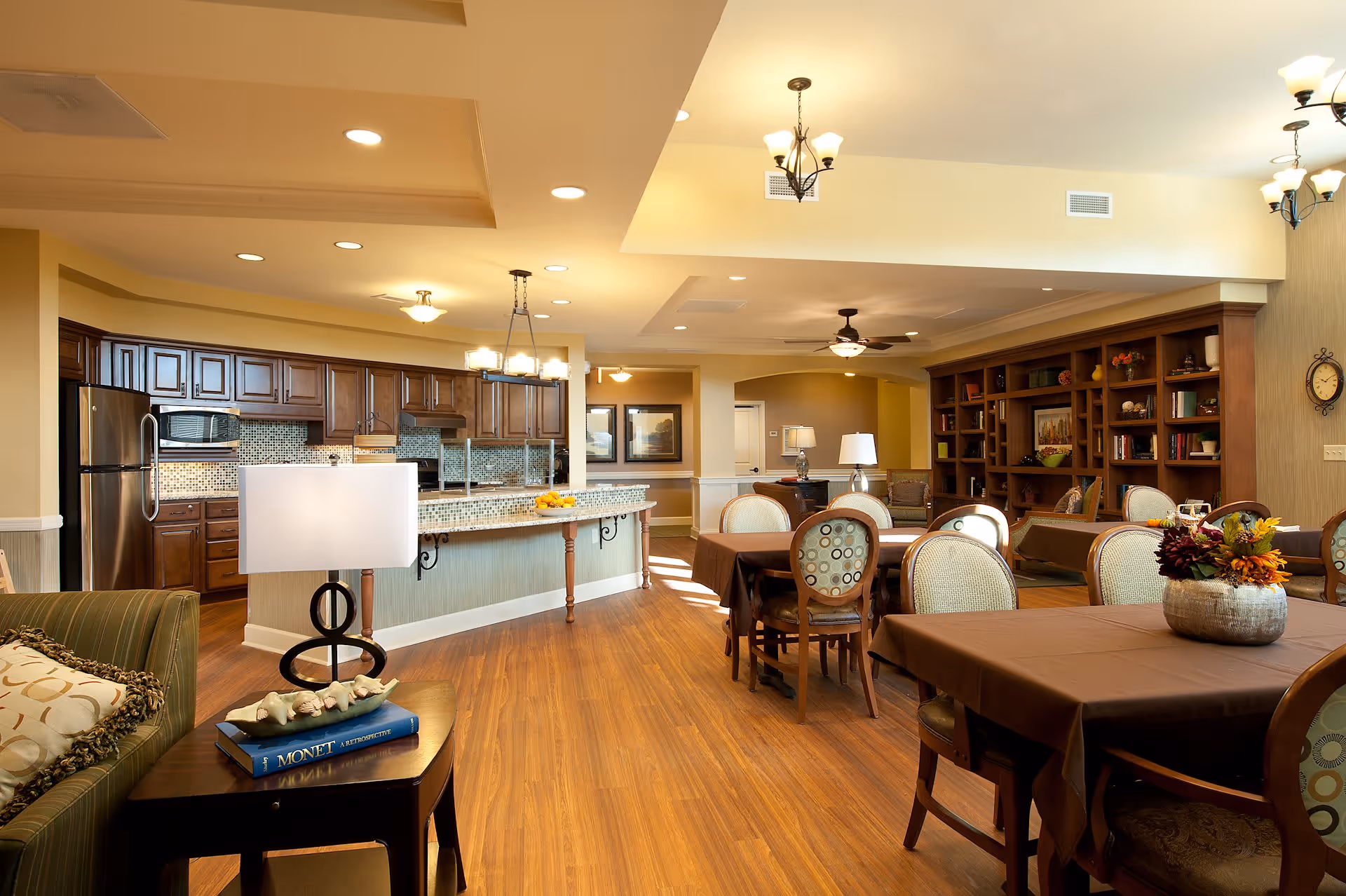 A spacious and well-lit common area in a senior living facility featuring a kitchen with dark wood cabinets and stainless steel appliances, a dining area with tables covered in brown tablecloths and chairs, and a living room area with a large wooden bookshelf filled with decorative items. The room has warm wood flooring, ceiling fans, and multiple light fixtures.