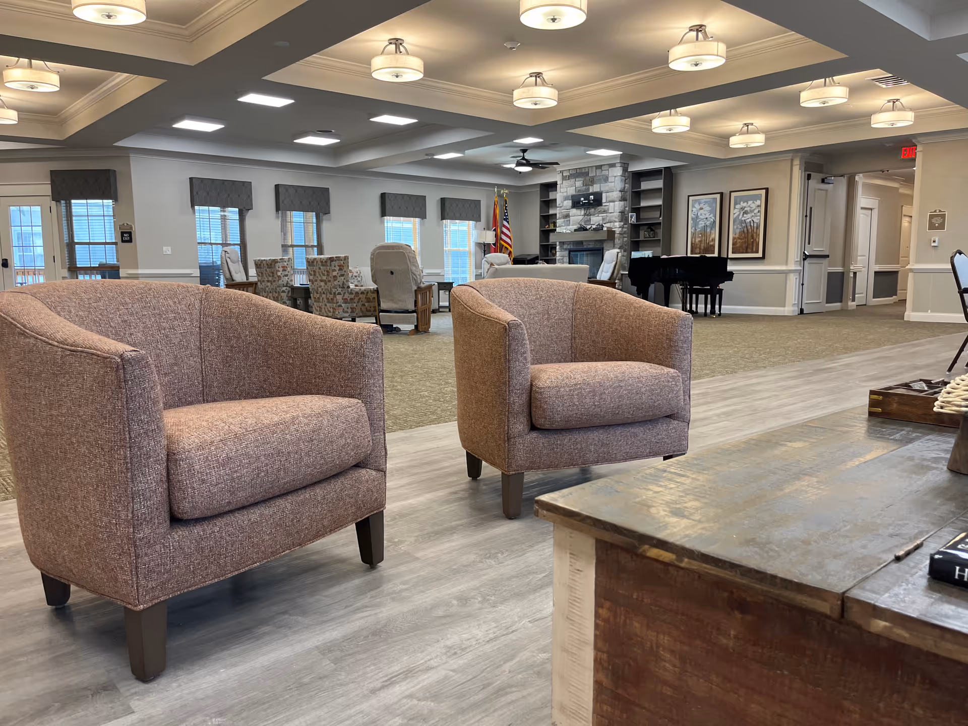 A spacious and well-lit common area in a senior living facility with two upholstered armchairs in the foreground, a wooden coffee table, multiple seating arrangements in the background, a stone fireplace, an American flag, and framed artwork on the walls.
