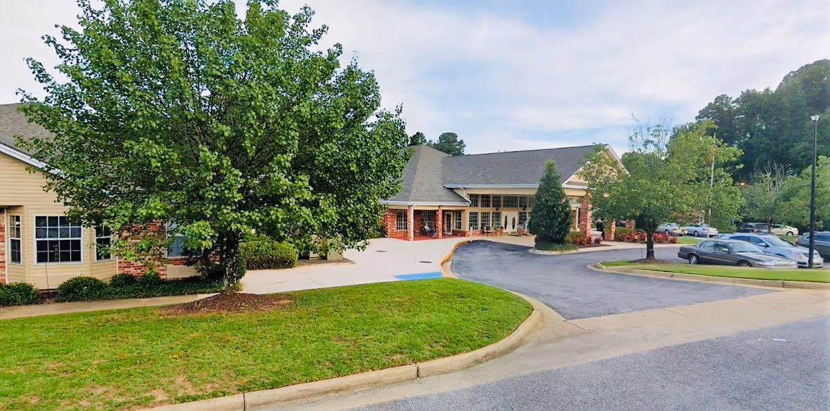 Exterior view of a senior living facility building with a driveway and parking area. The building has a combination of brick and light yellow siding, with large windows and a covered entrance. There are green trees and well-maintained grass surrounding the driveway and parking lot.