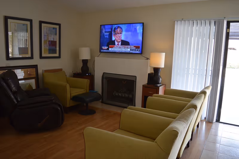 A cozy living room with several green armchairs and a recliner facing a wall-mounted TV above a fireplace, next to sliding glass doors with vertical blinds.