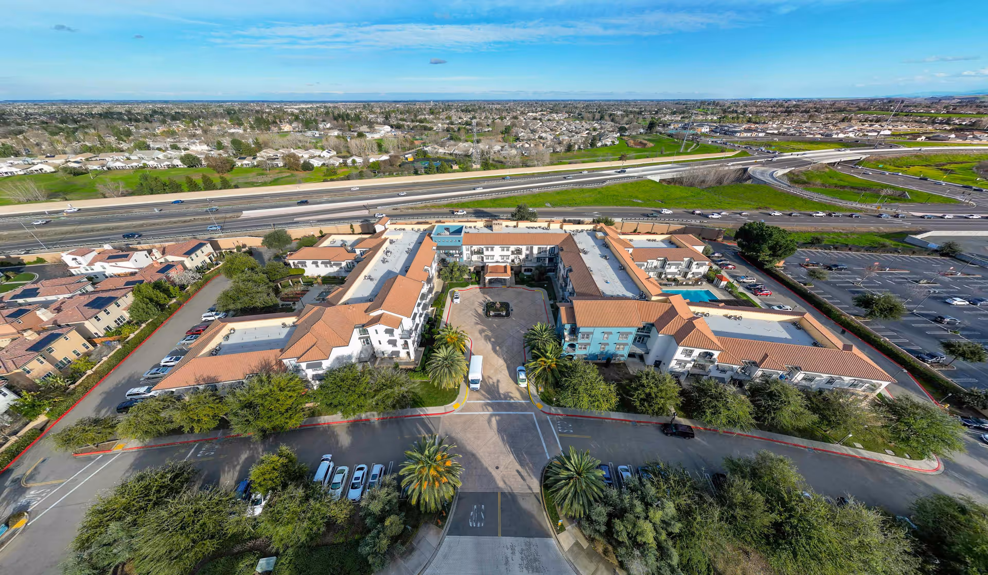 Aerial view of Cogir of Brentwood senior living facility showing a large U-shaped building with terracotta roofs surrounded by trees, parking lots, and nearby highways under a blue sky.