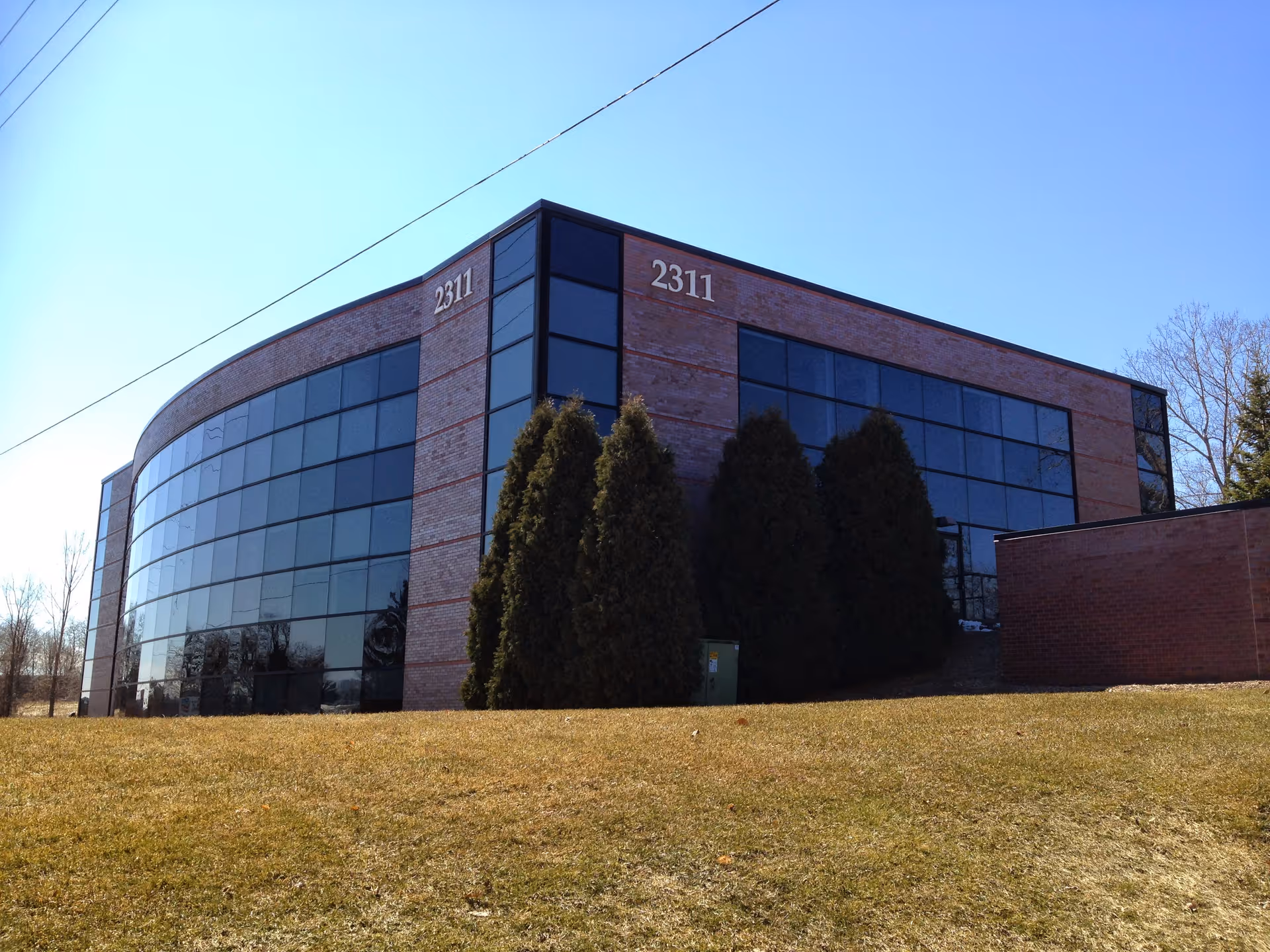 Curved glass-front building with brick accents sits on a grassy, sunlit lot.