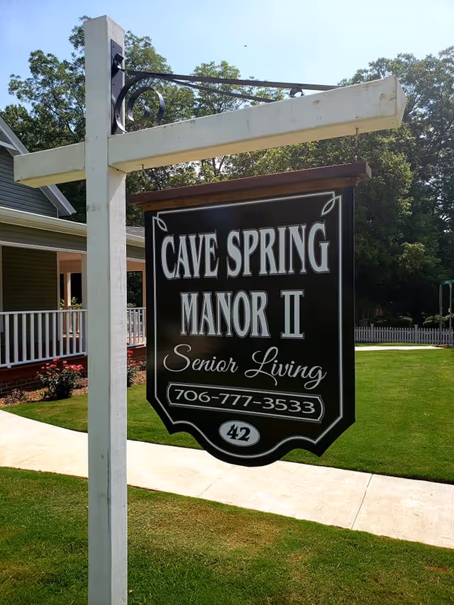A hanging black sign reads Cave Spring Manor II Senior Living beside a white support post.