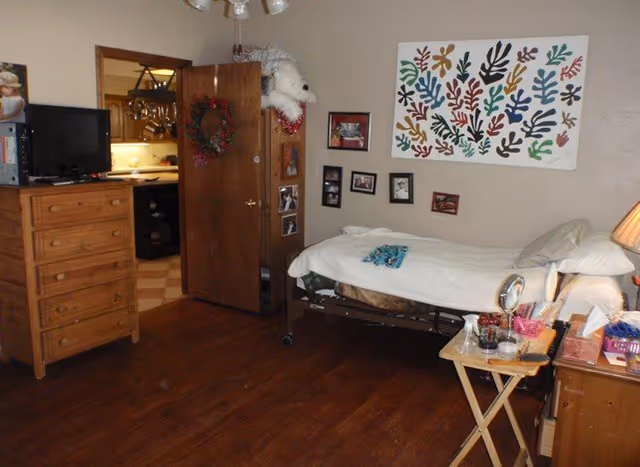 A resident bedroom with a hospital bed, dresser, TV, photos, and open doorway to a kitchen.