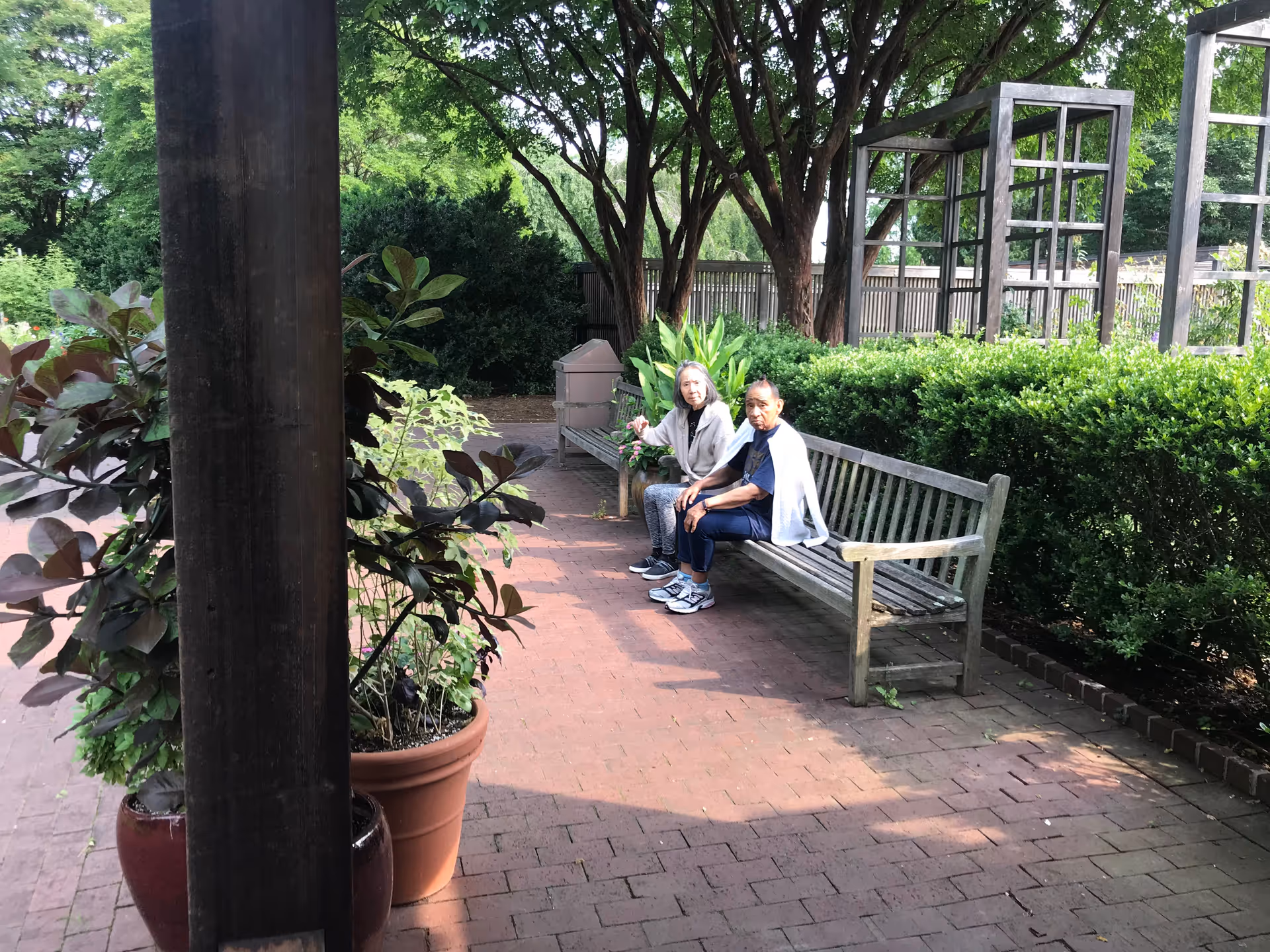 Two elderly women sitting on a wooden bench in a garden area with brick pavement, surrounded by green bushes, trees, and potted plants. There is a wooden pergola structure to the right and a wooden post with plants in pots to the left.