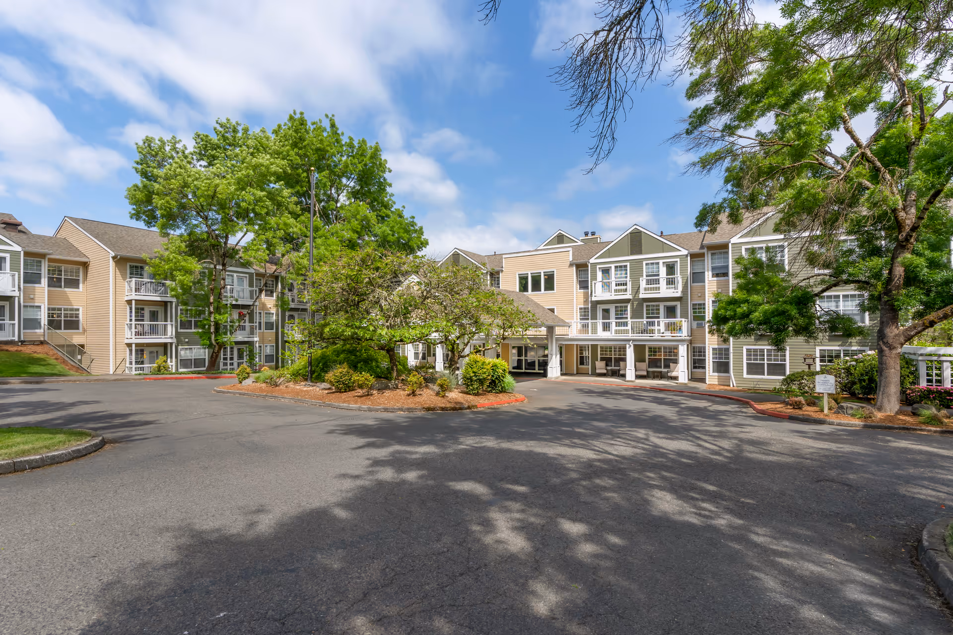 Front exterior of a multi-story senior living building with a circular driveway, landscaped island, and balconies under a partly cloudy sky.