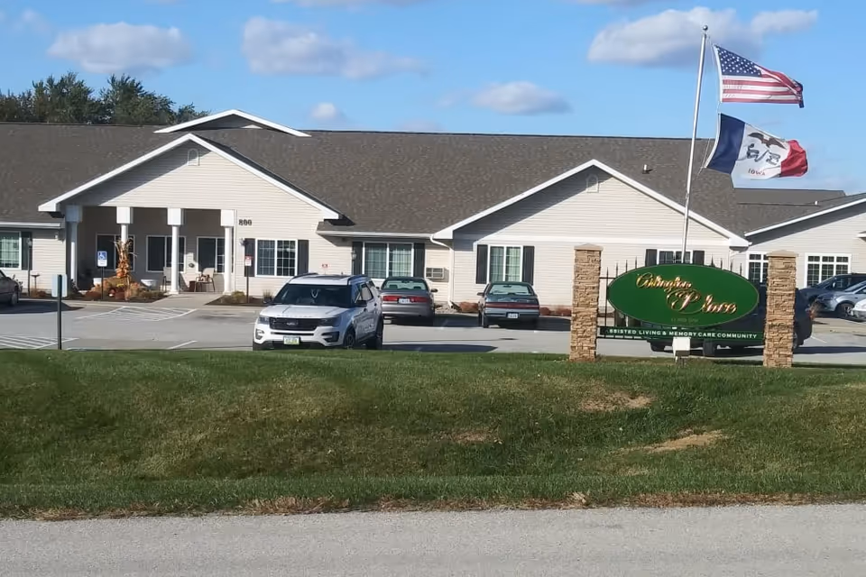 Front exterior of a single-story senior living building with a parking lot, flags, and a green entrance sign.