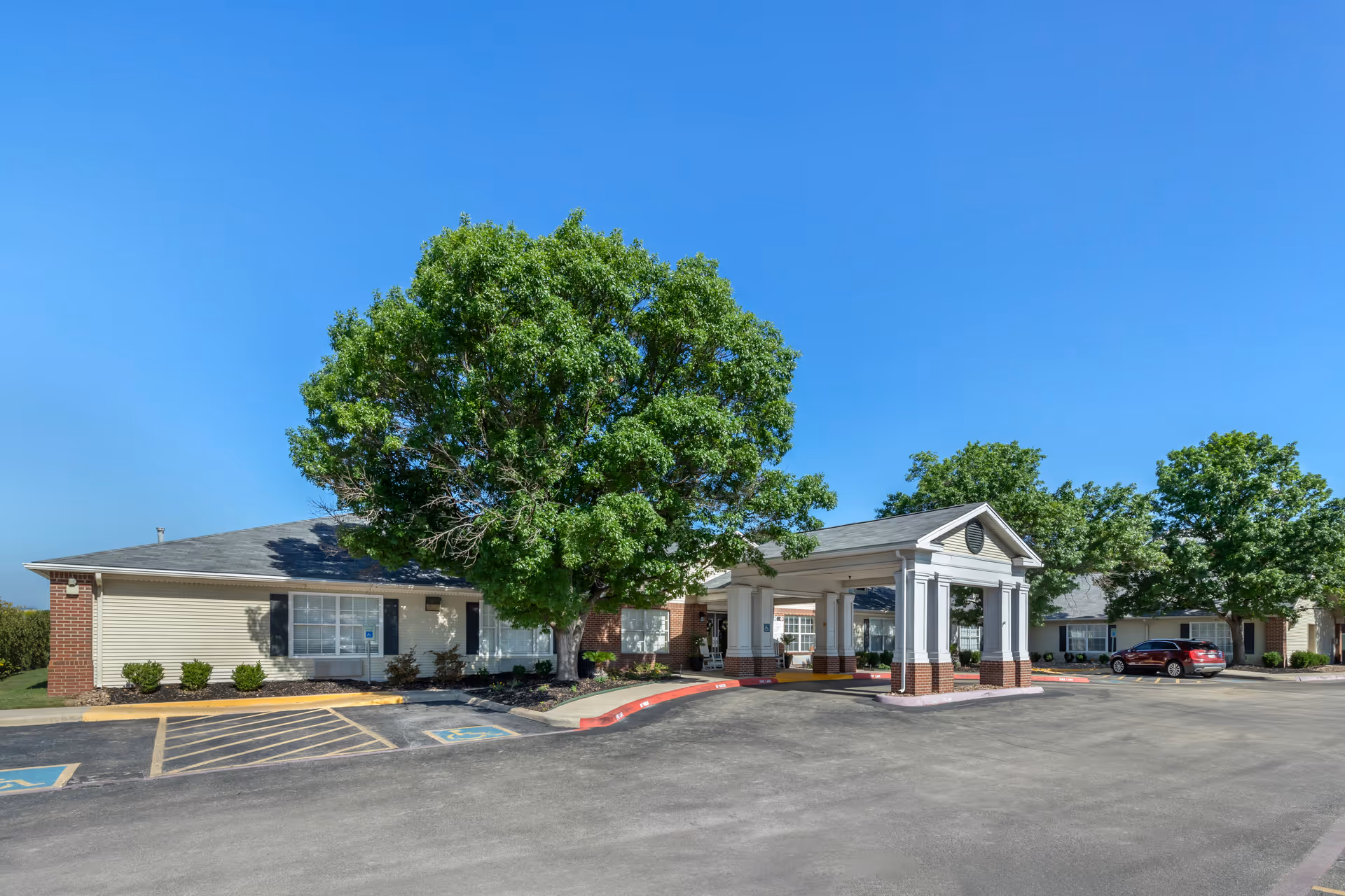 Front entrance of a single-story senior living building with a covered portico, large tree, and parking area under a clear blue sky.