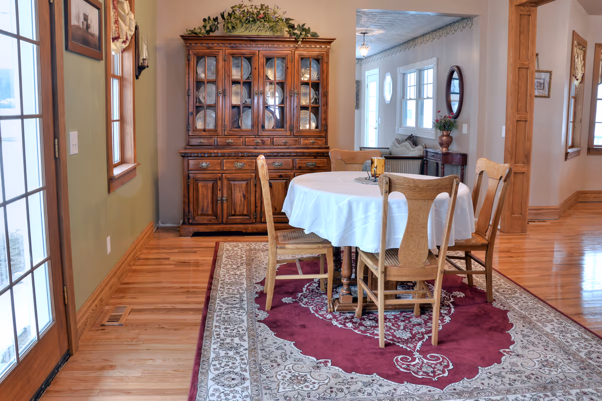 Dining room with a round table, white cloth, wooden chairs, and a china cabinet.