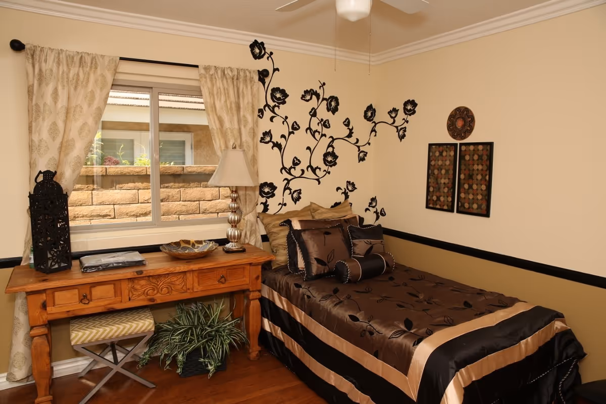 Cozy bedroom in a senior living suite with bed, desk, lamp, and wall decor.