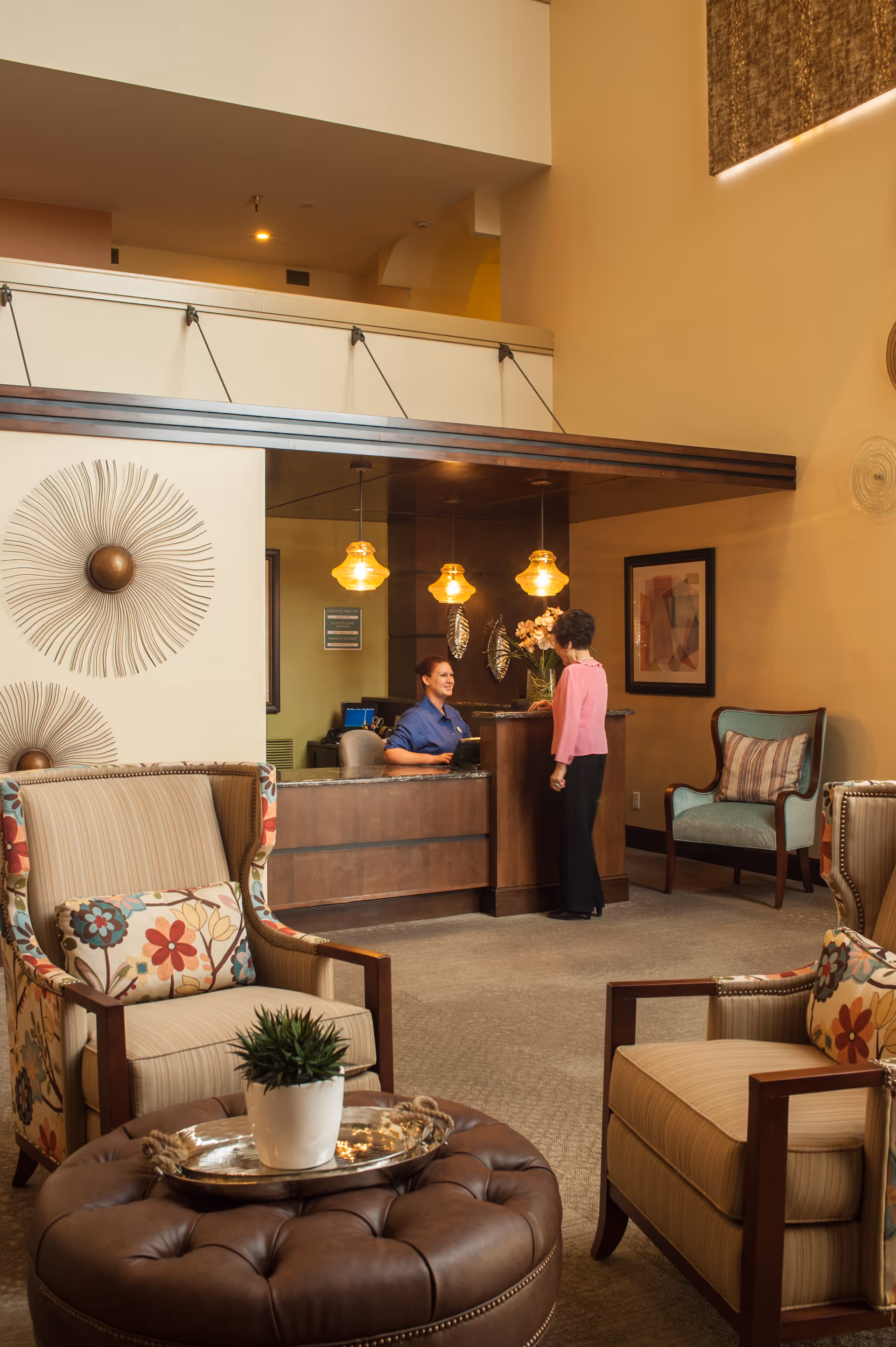 A cozy reception area in a senior living facility with a receptionist sitting behind a wooden desk talking to a visitor. The room features comfortable armchairs with floral and striped cushions, a round leather ottoman with a silver tray and a potted plant, warm lighting from hanging pendant lamps, and decorative wall art.