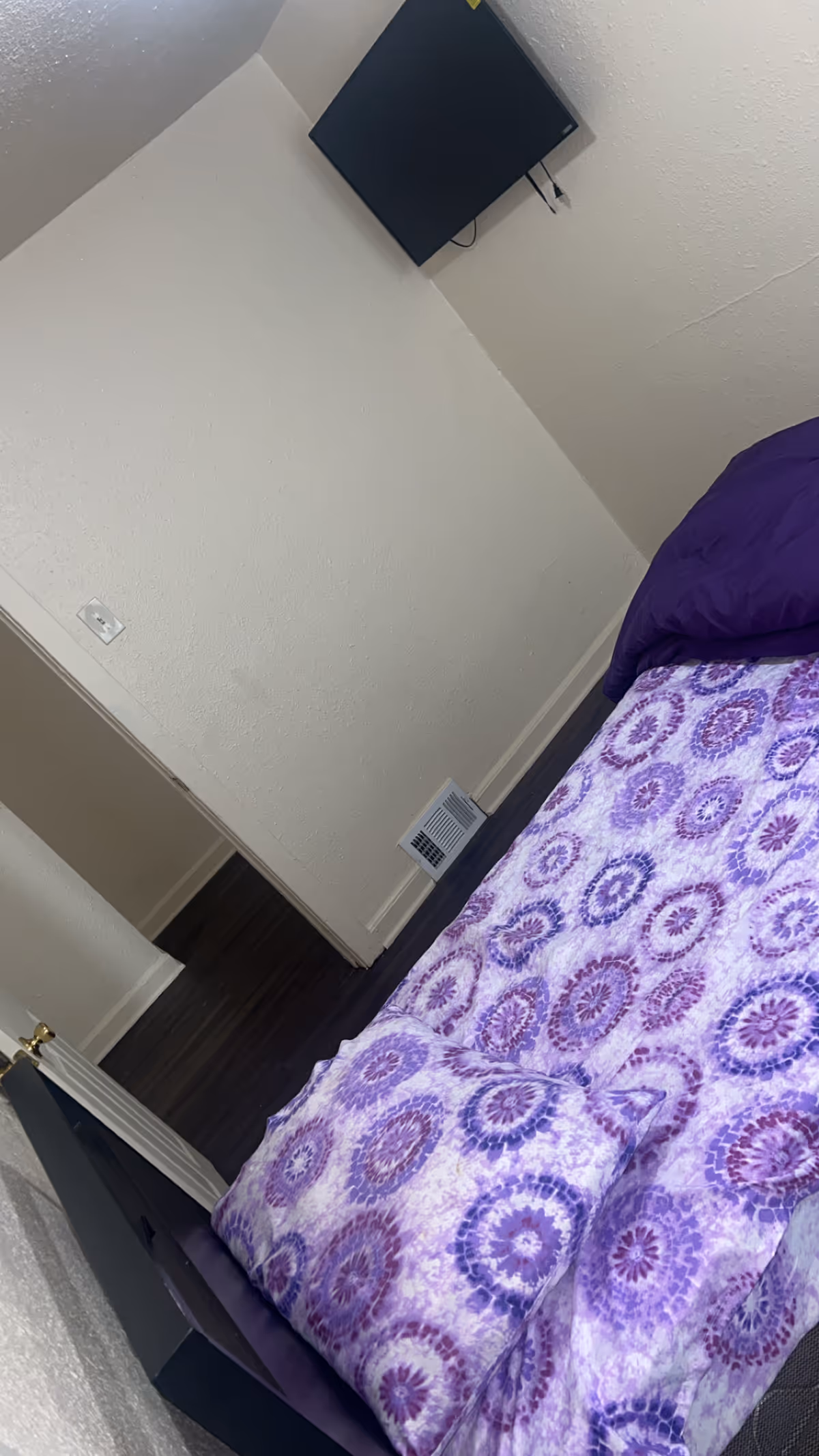 Bedroom with a purple patterned bedspread, wall-mounted TV, and a floor vent.