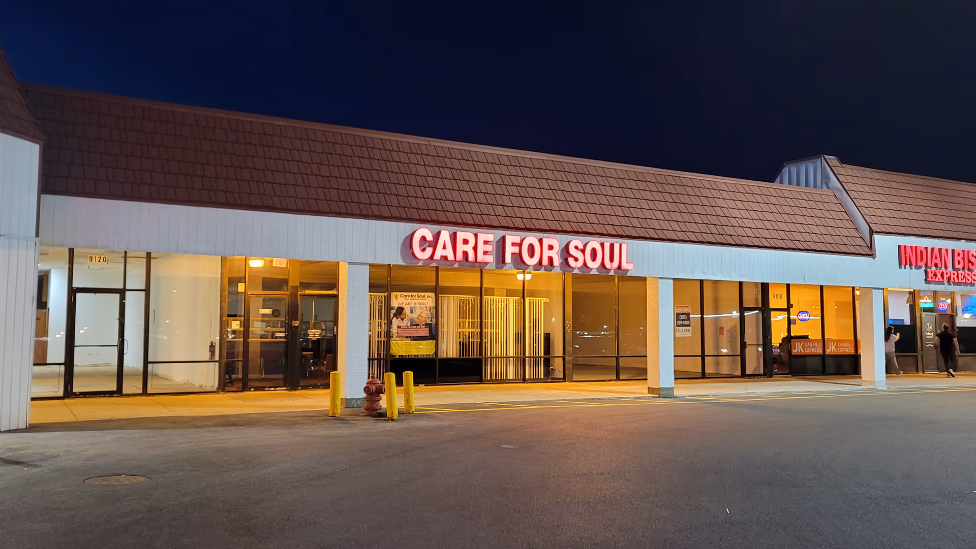 Night exterior of a strip mall with 'Care For Soul' storefront beside Indian Bistro.