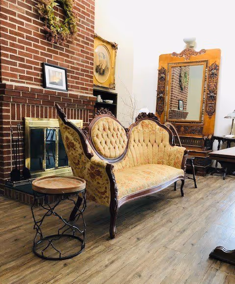A cozy interior space featuring a vintage tufted sofa with wooden frame, positioned near a brick fireplace with a brass screen. Above the fireplace hangs a wreath and framed pictures. To the right, there is an ornate wooden mirror and a small side table with a lamp, all set on wooden flooring.