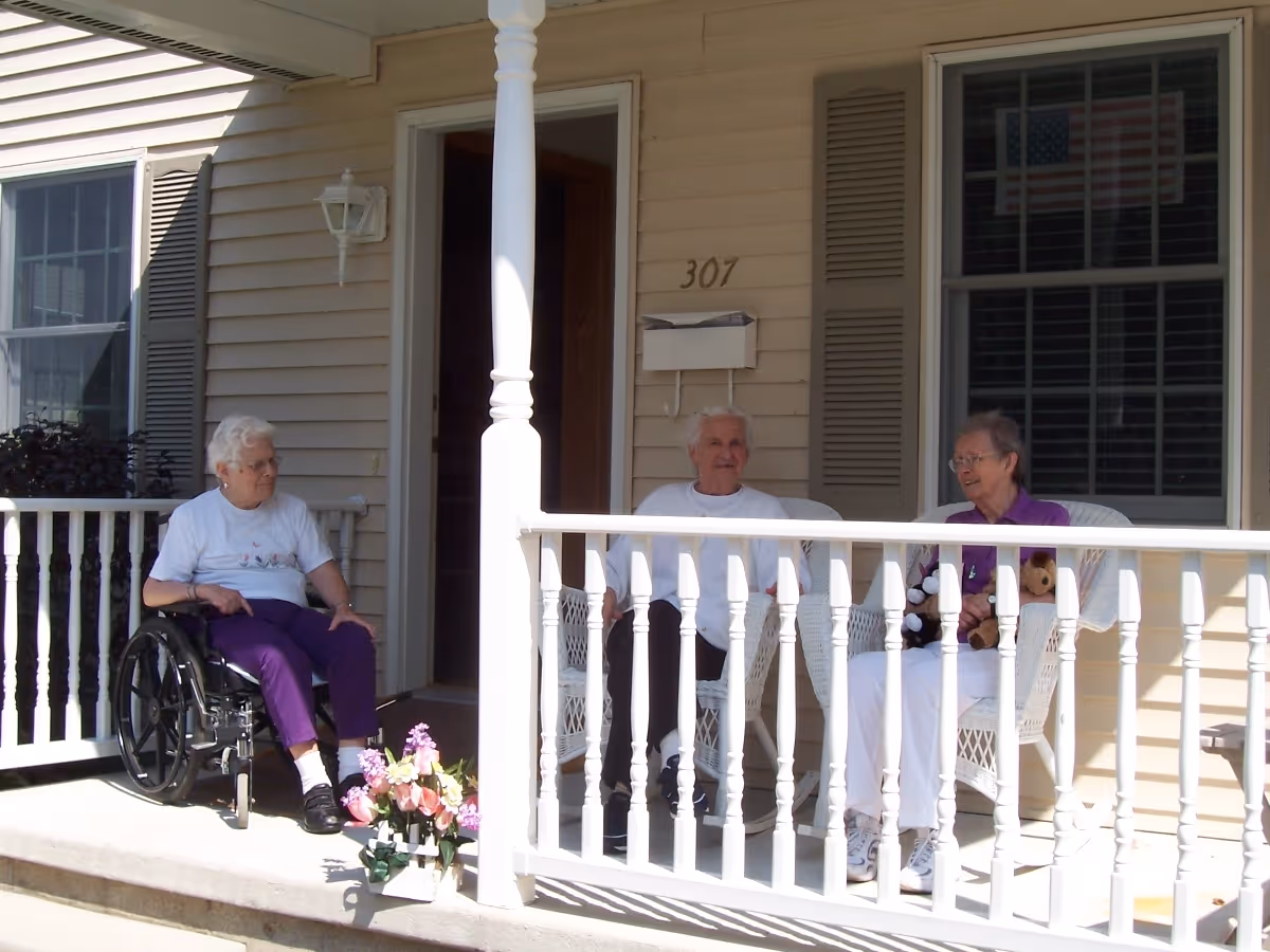 Three seniors, one in a wheelchair, sit on a porch outside a beige home.