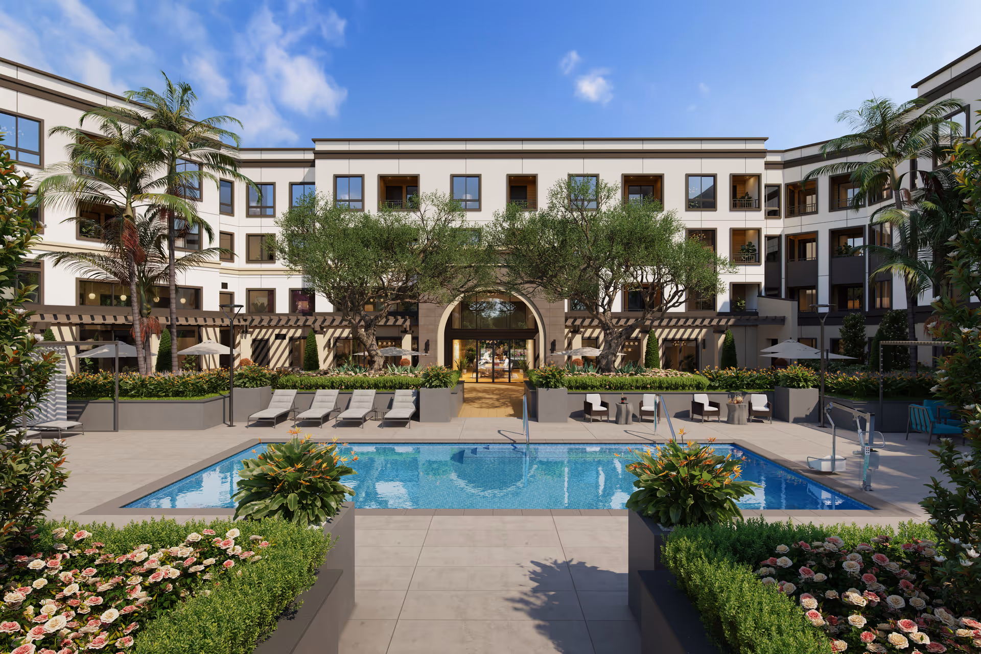 Outdoor courtyard area of a senior living facility with a swimming pool surrounded by lounge chairs and greenery, including palm trees and flowering plants. The background shows a multi-story building with balconies and large windows under a blue sky with some clouds.