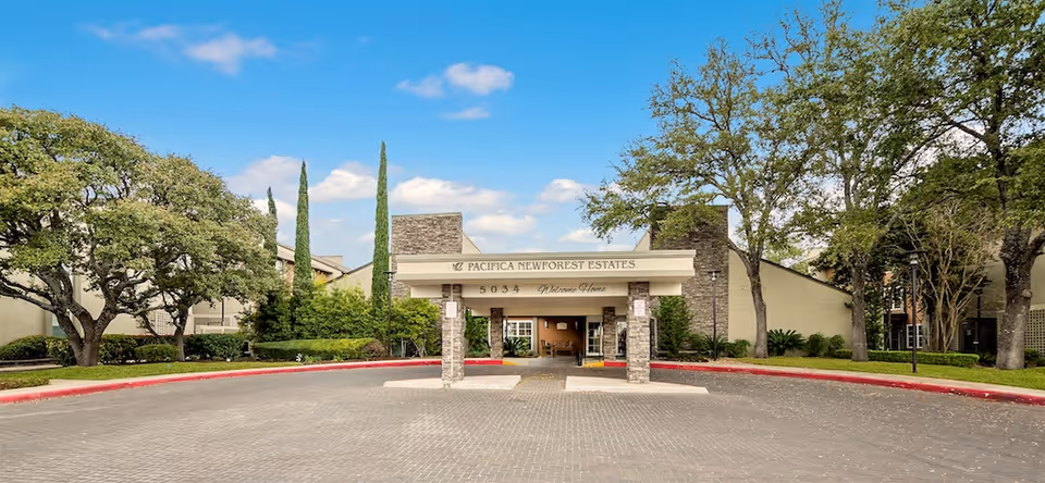 Front entrance canopy and circular driveway of New Forest Haven senior living facility flanked by trees under a blue sky.