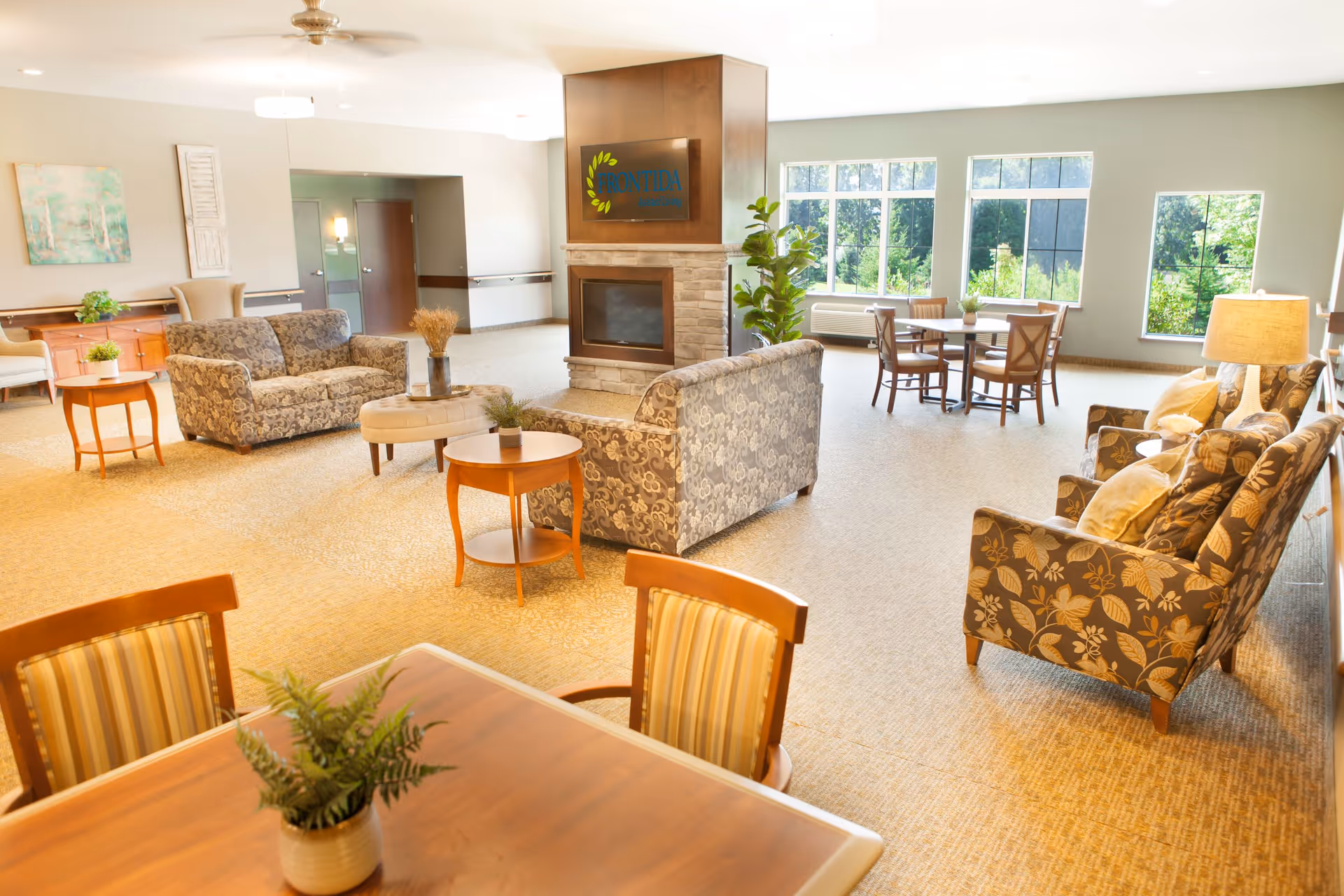 A spacious and well-lit common area in a senior living facility featuring multiple seating arrangements with patterned sofas and armchairs, a central fireplace with a Frontida of Germantown sign above it, large windows letting in natural light, and several small tables with chairs and potted plants.