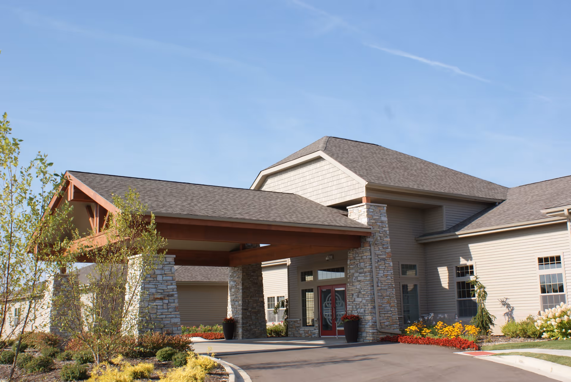 Exterior view of Cambrian Assisted Living and Memory Care South Lyon building entrance with a covered driveway supported by stone pillars, surrounded by landscaped plants and flowers under a clear blue sky.