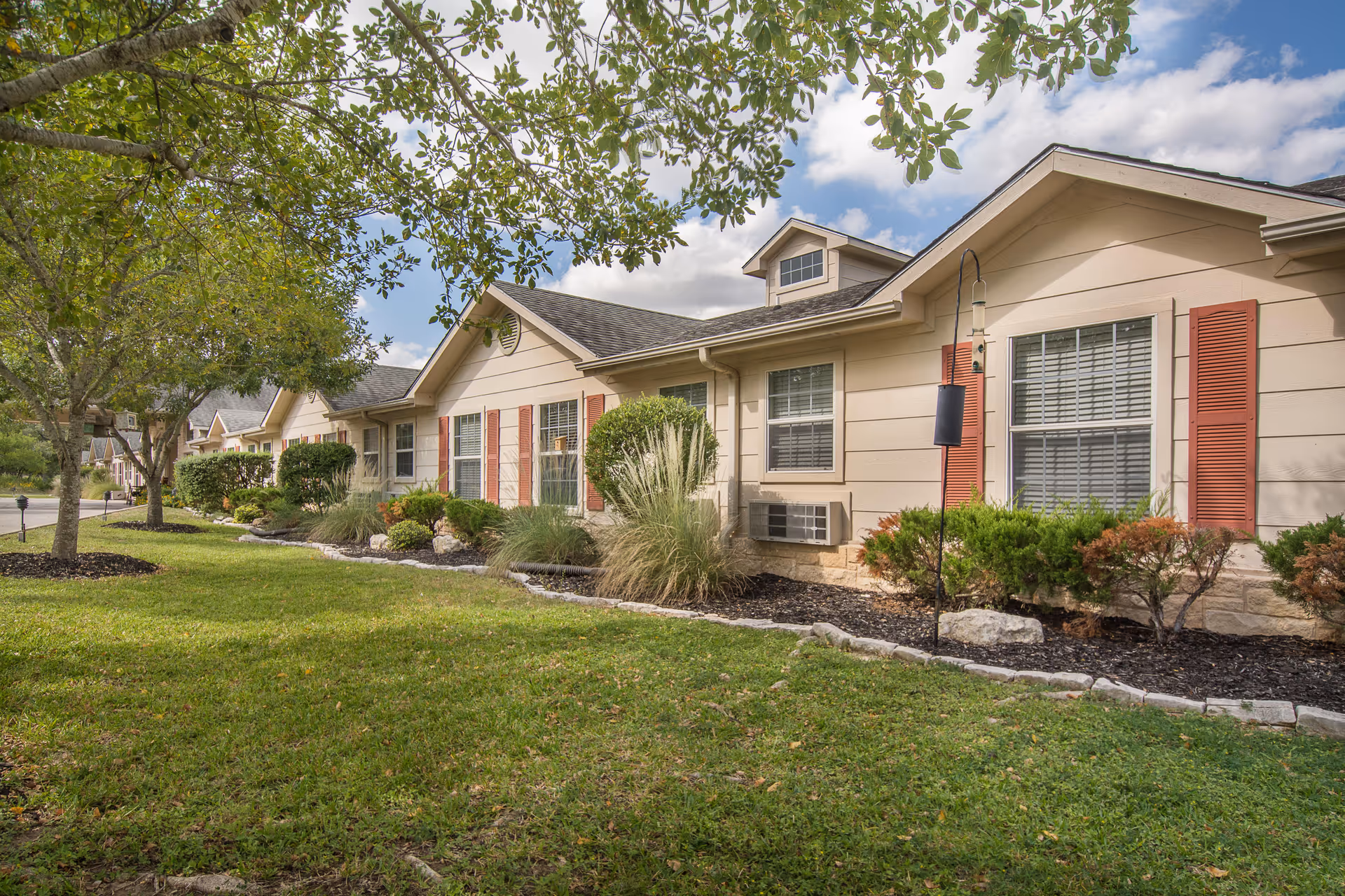 Exterior view of a single-story residential building with beige siding and red shutters, surrounded by green grass, shrubs, and trees under a partly cloudy sky.
