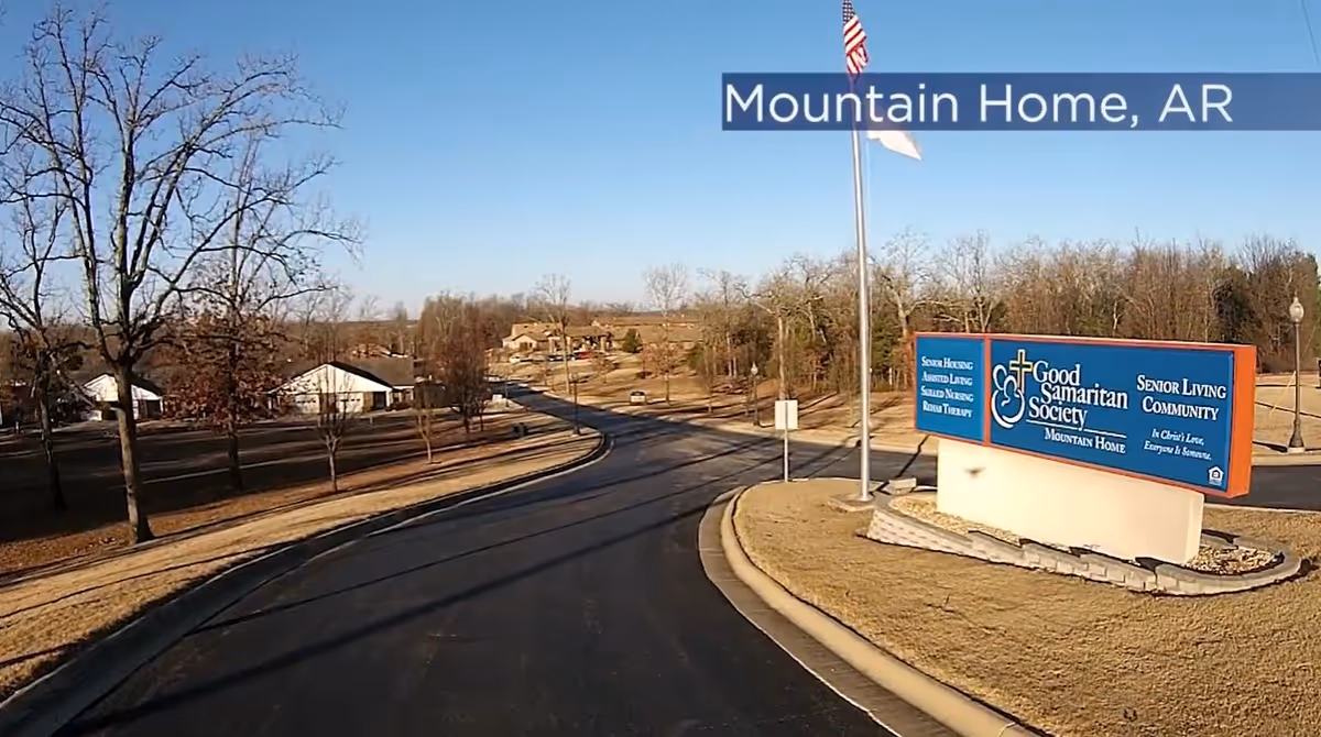 View of the entrance road to a senior living community with a large sign that reads 'Good Samaritan Society Mountain Home Senior Living Community' and an American flag on a flagpole. Trees and buildings are visible in the background under a clear blue sky.