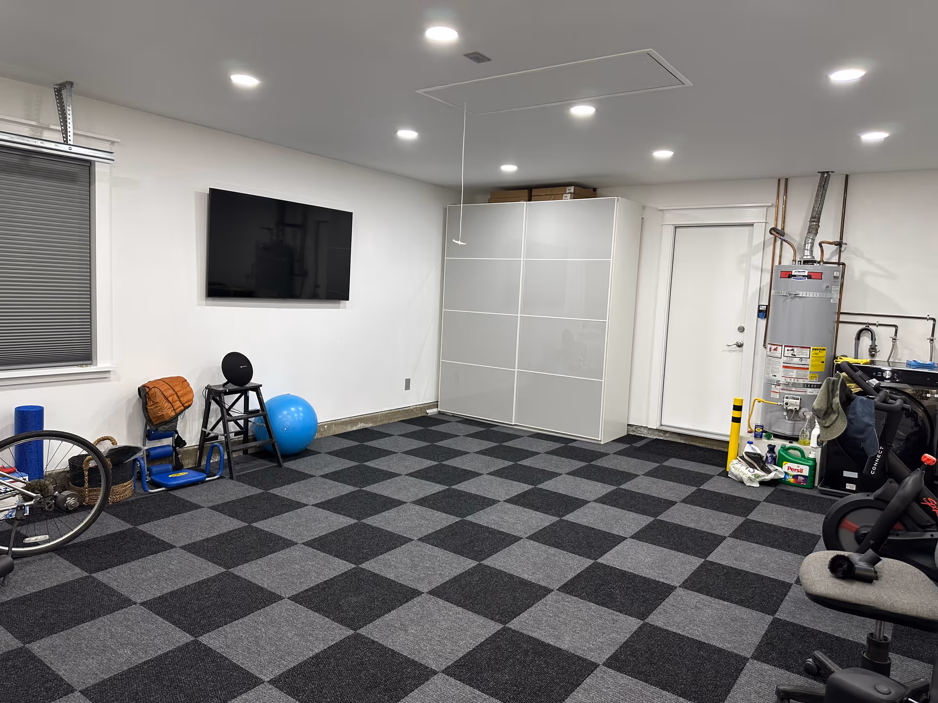 A spacious room with checkered black and gray carpet tiles, a wall-mounted flat screen TV, exercise equipment including a blue exercise ball and a stationary bike, a white cabinet, and a water heater near a white door. The room has white walls and ceiling with recessed lighting.