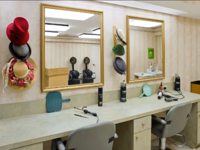 Interior view of a salon area with two chairs in front of a long countertop. Two large mirrors with gold frames are mounted on the wall above the counter. Various hair styling tools and products are placed on the countertop, and several hats are hanging on the wall between the mirrors.