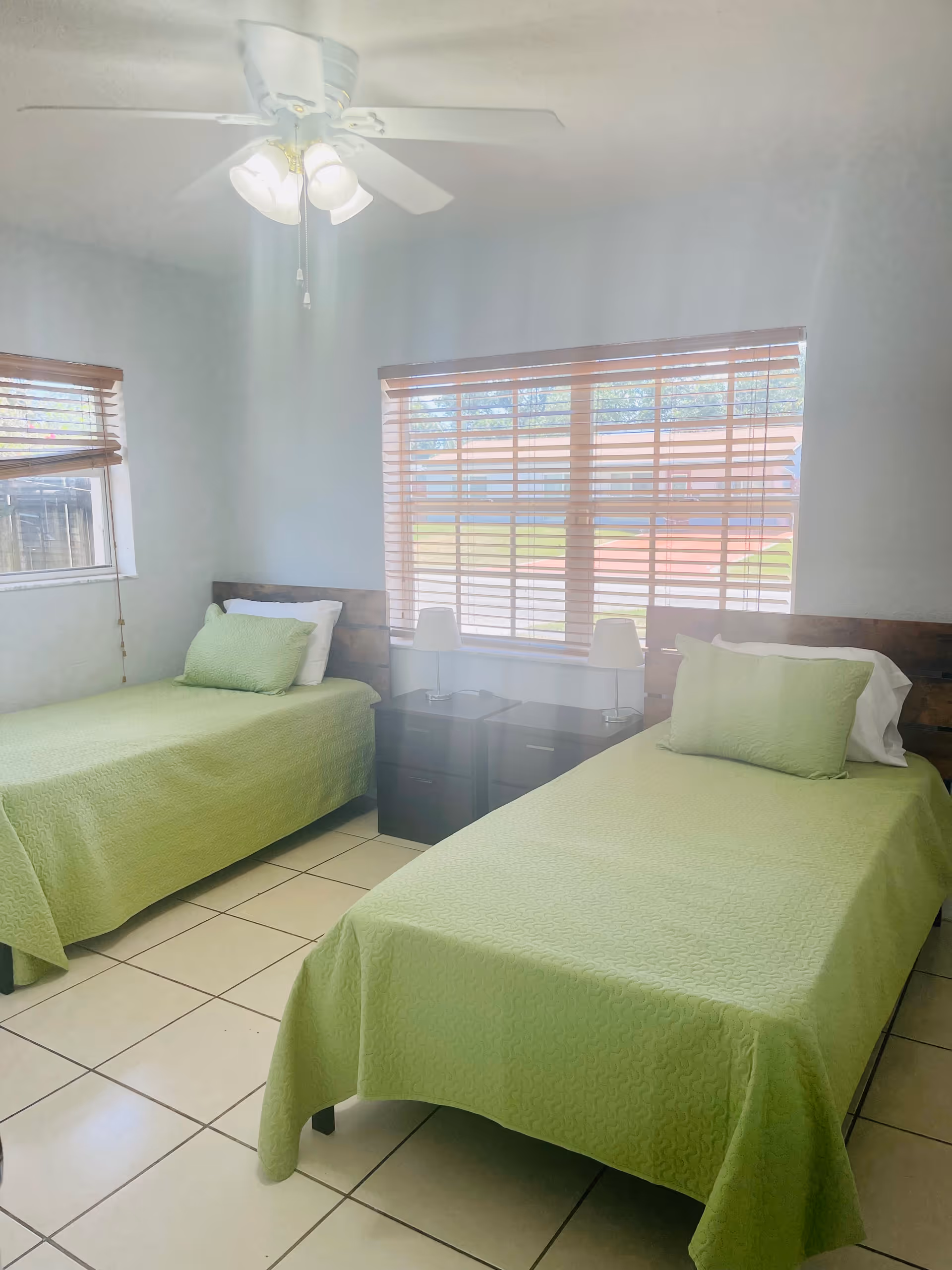 Two single beds with green bedding, a shared nightstand, blinds, and a ceiling fan.