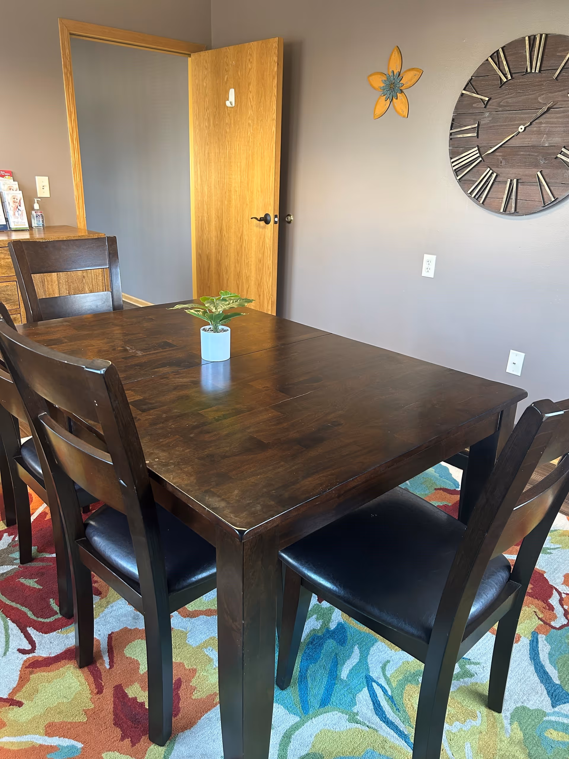 A dining area with a dark wooden table and four matching chairs with black cushioned seats. A small potted plant is placed in the center of the table. The room has a colorful patterned rug underneath the table and chairs. On the wall, there is a large round wooden clock and a decorative yellow flower. A wooden door is open, revealing a hallway with light-colored walls.