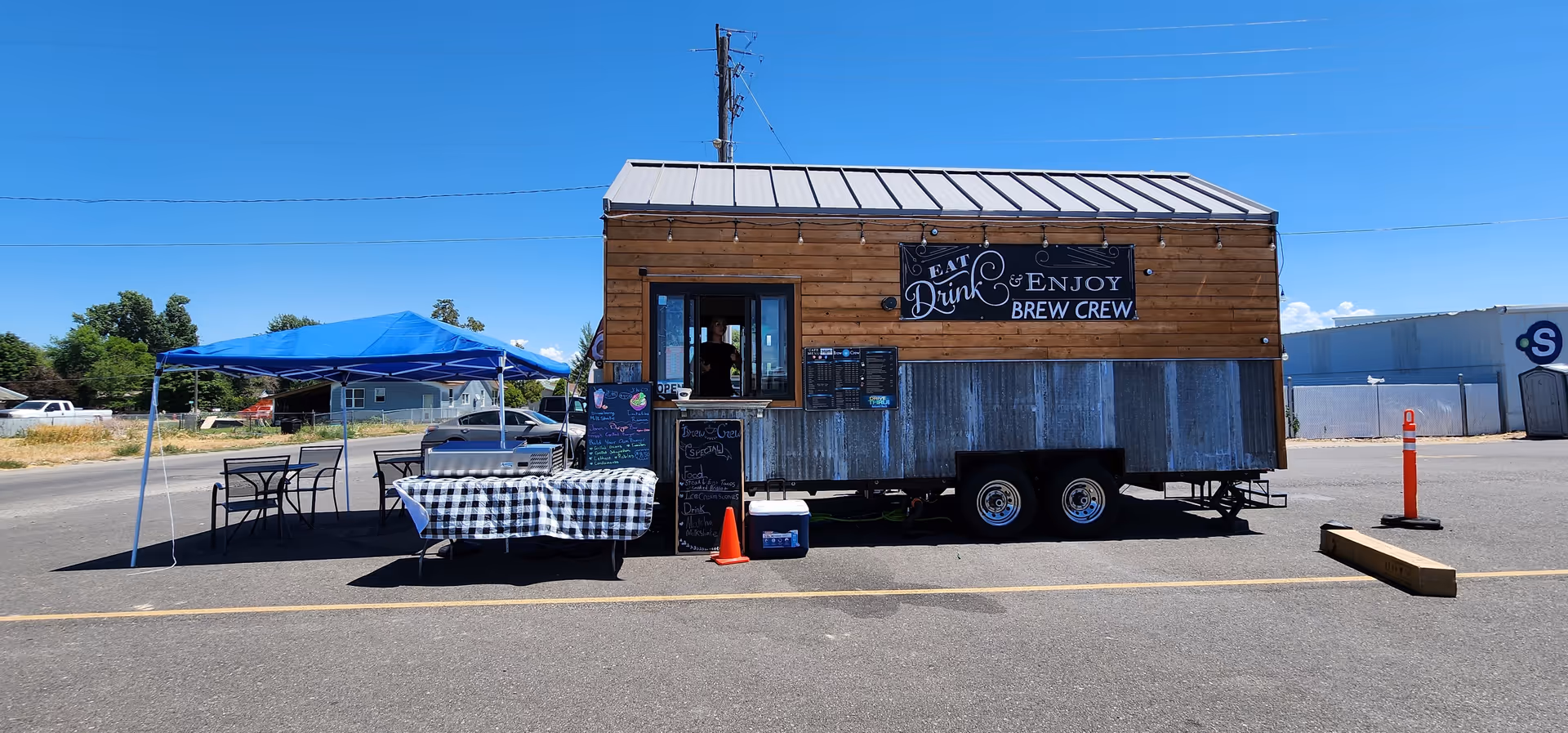 A wooden food-truck trailer with a blue canopy and checkered tablecloths outside.