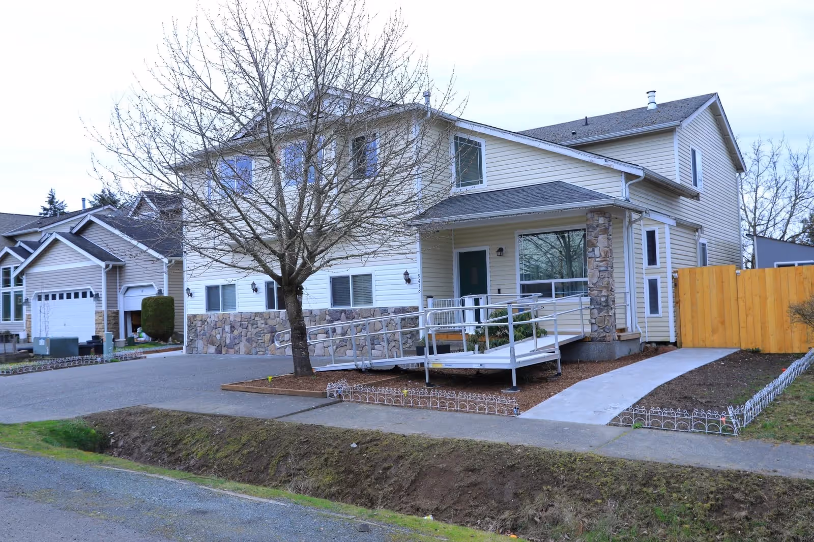 Two story beige adult family home with stone facade, ramped entrance, and wooden fence.