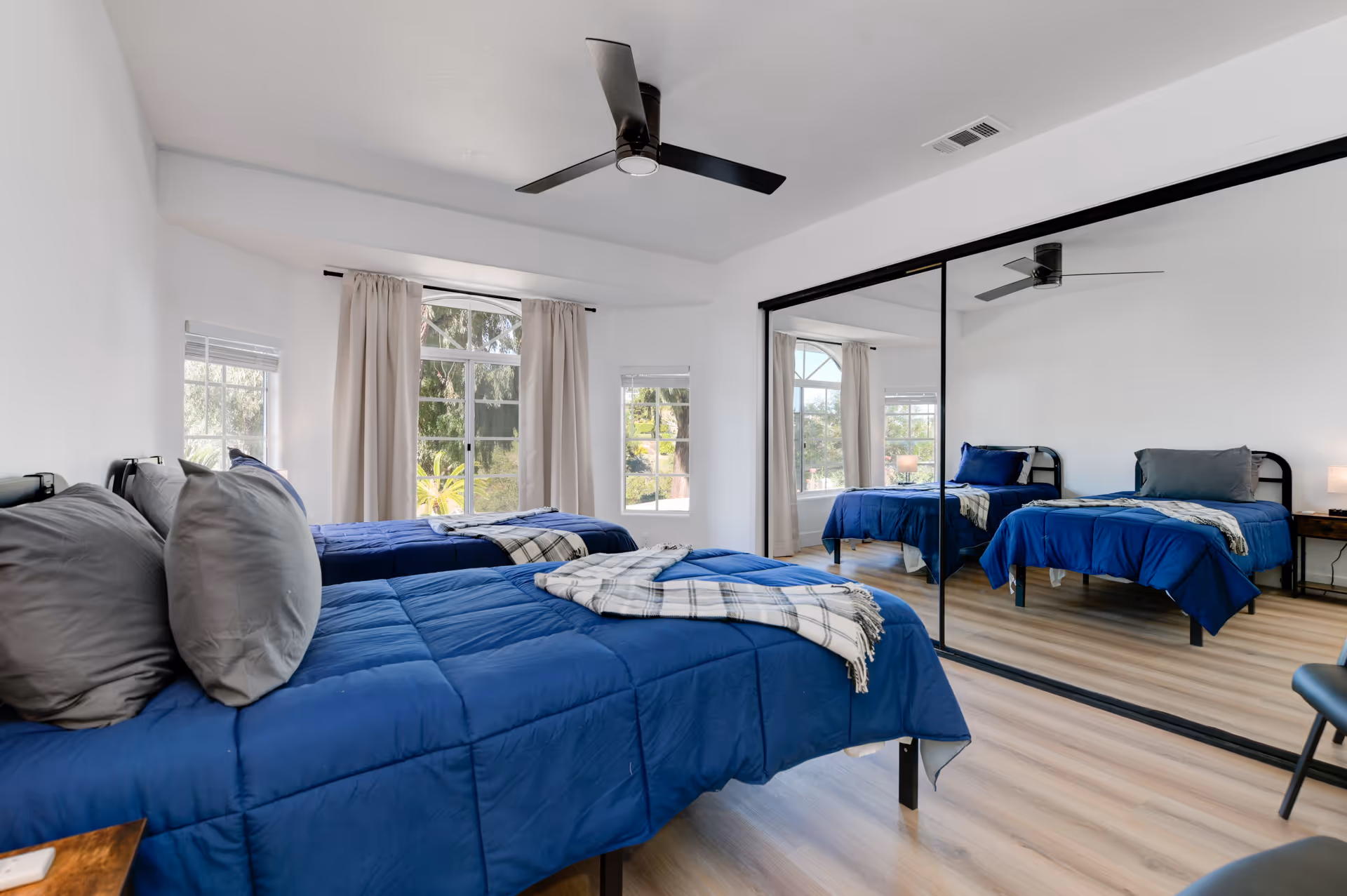 Bright shared bedroom with two blue beds, mirrored closet, and large windows.
