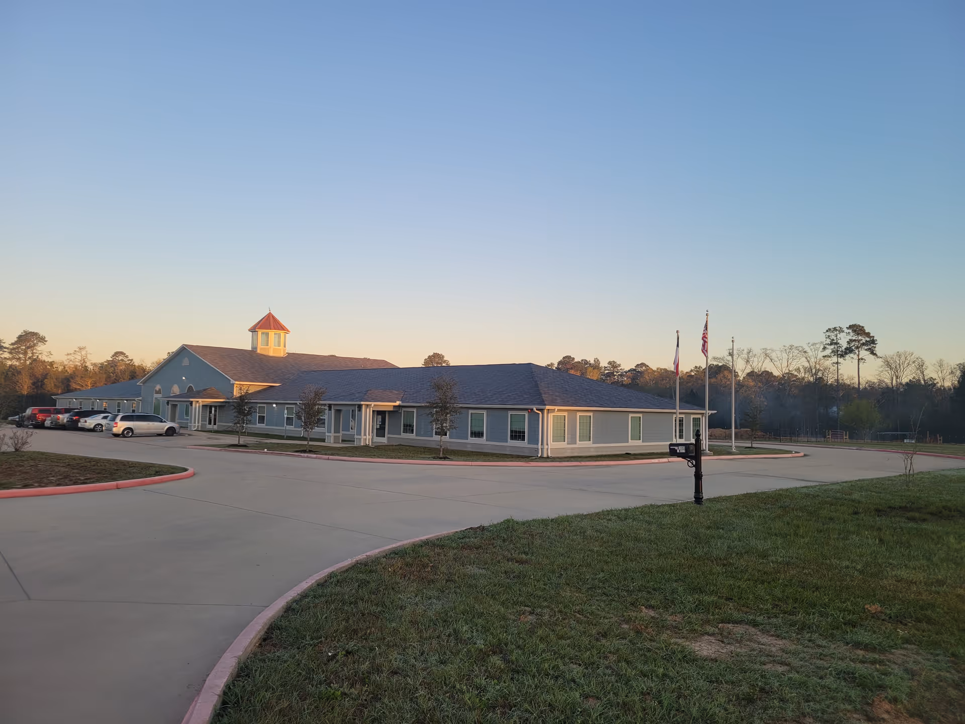 Single-story Jasper Harbor Hospice of East Texas facility with curved driveway, parking, and flagpoles at sunset.