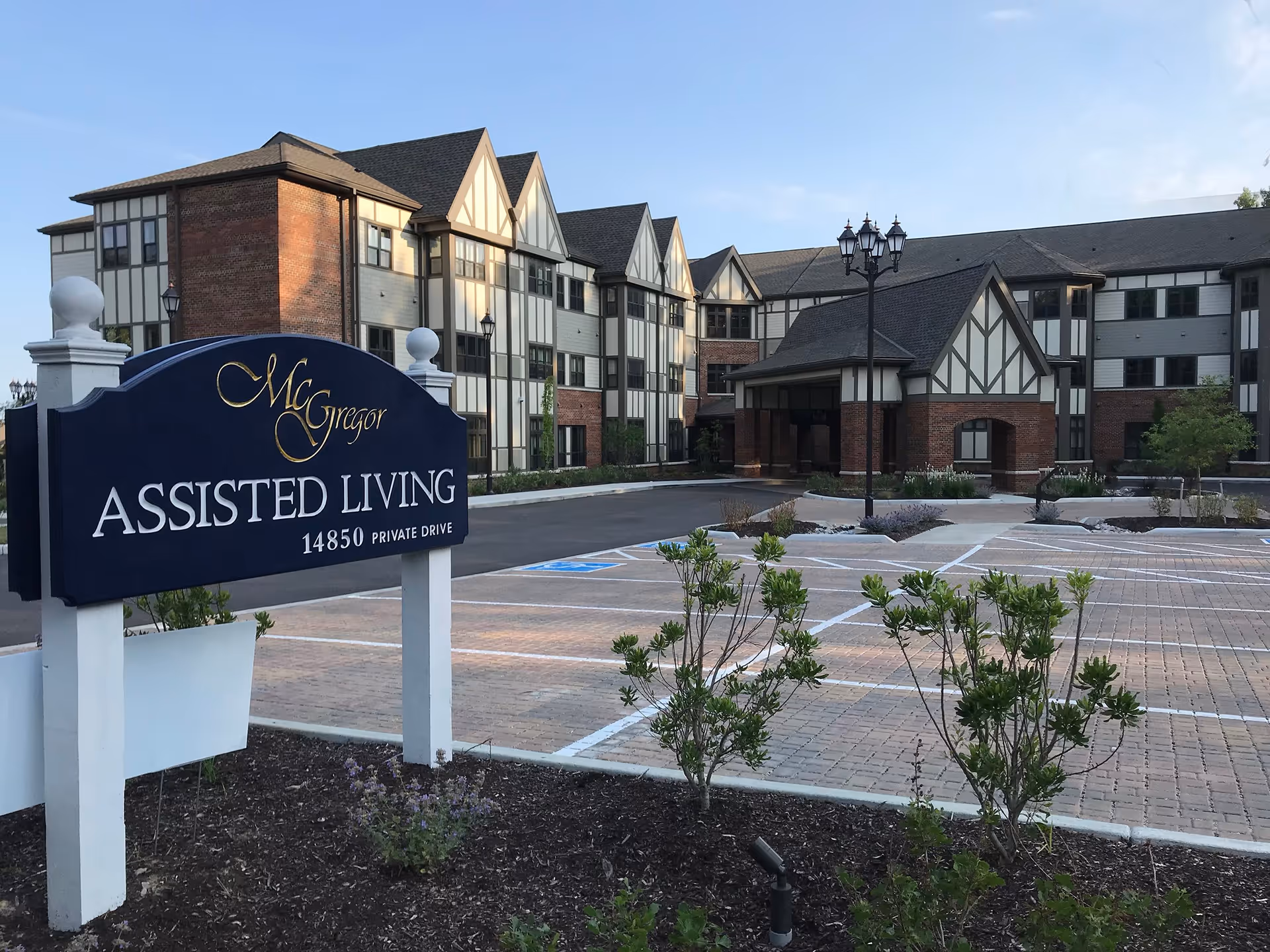 Exterior view of McGregor Assisted Living facility showing a large multi-story building with Tudor-style architecture, a parking lot with marked spaces, and a sign in the foreground that reads 'McGregor Assisted Living 14850 Private Drive'.