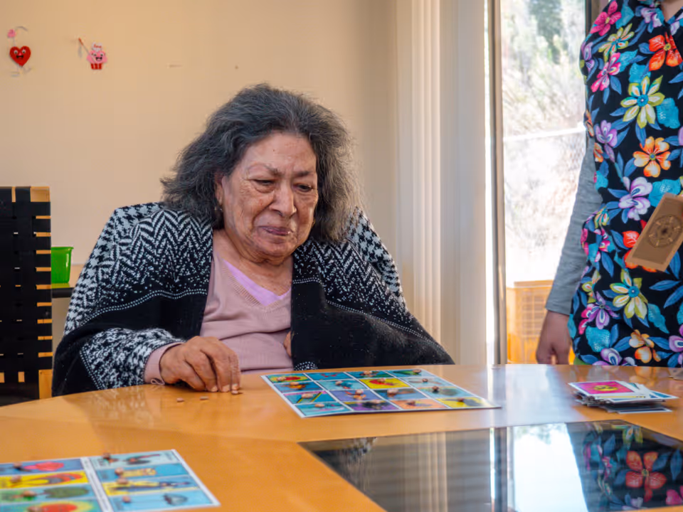 An elderly woman sitting at a wooden table playing a game with colorful cards, wrapped in a black and white patterned shawl. Another person wearing a floral apron is standing nearby. The room has beige walls and a large window letting in natural light.