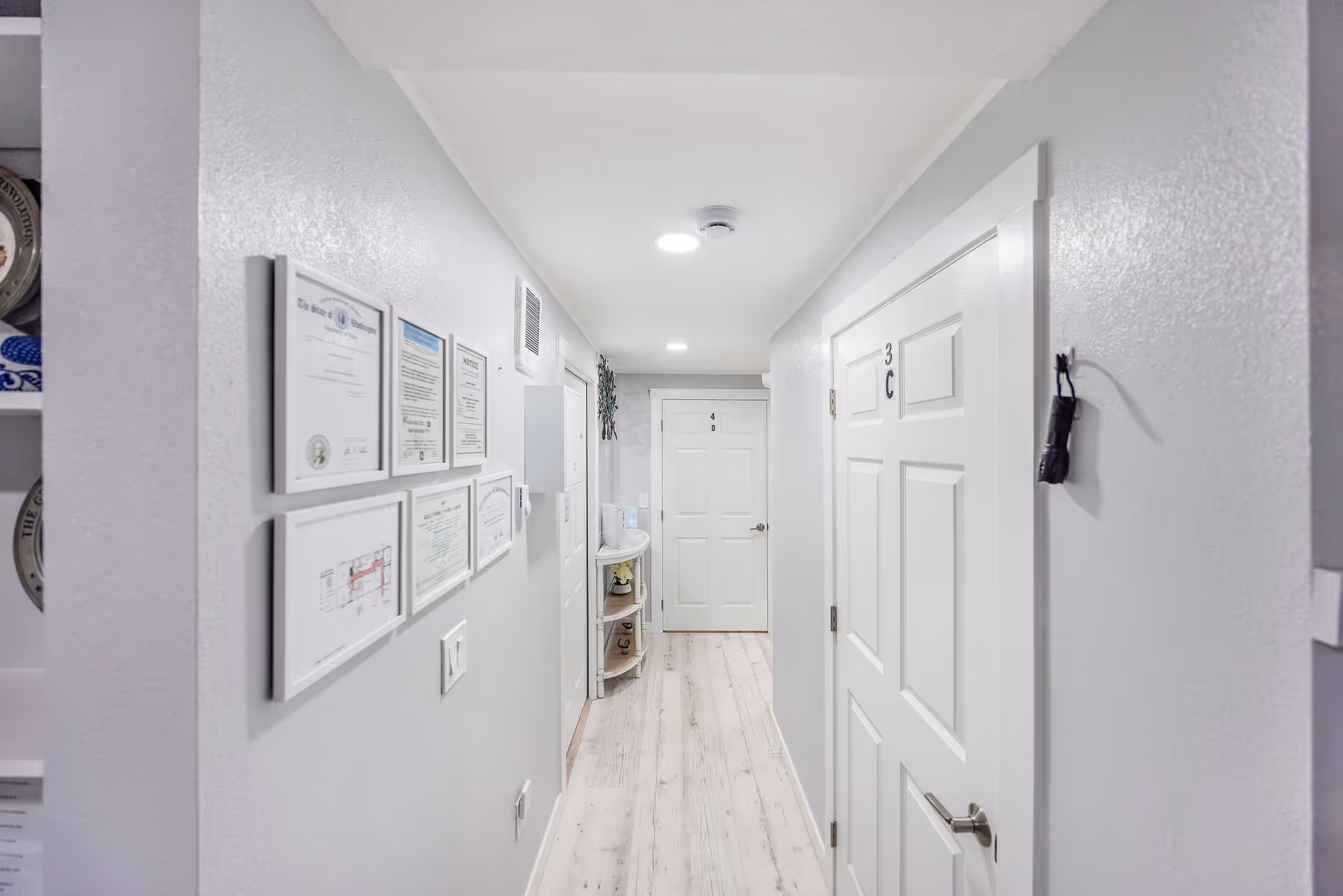 Bright hallway in a senior living facility with doors, framed certificates, and a small shelf.